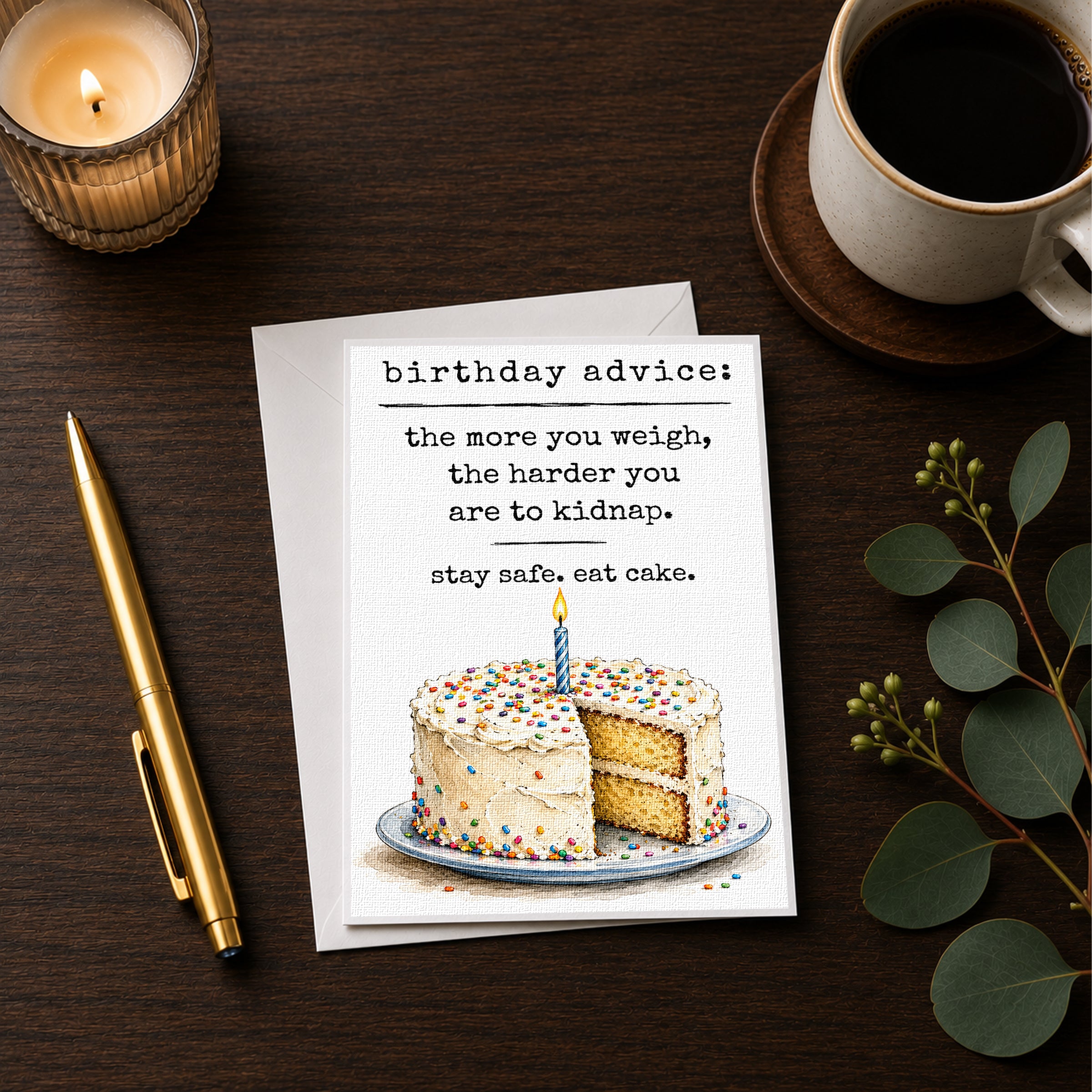 a birthday card with a cake and a candle, accompanied by a pen, a candle, and a cup of coffee on a wooden surface.