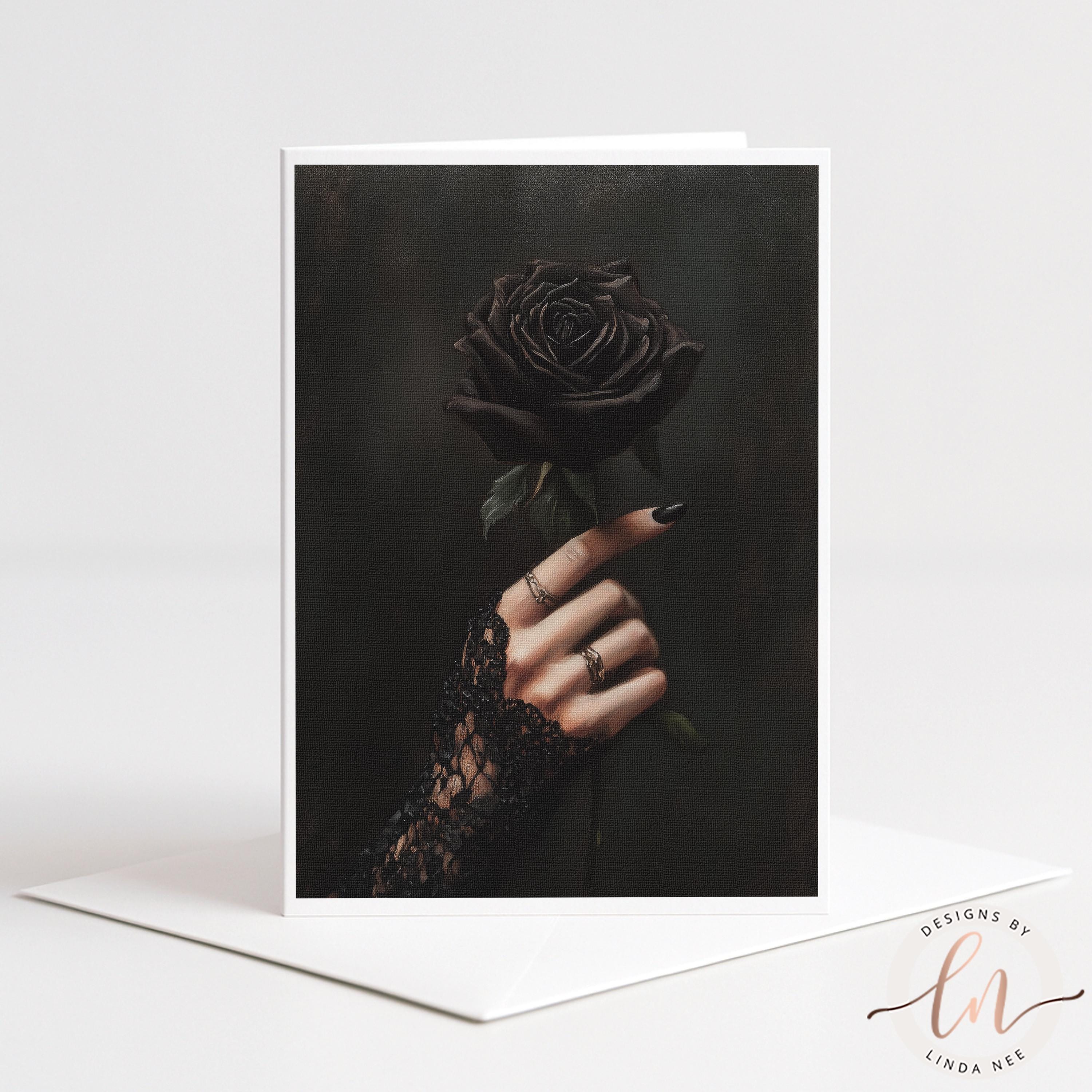 A vertical greeting card with a white border stands upright on a white envelope against a light gray background. The card features a dark, moody photograph of a hand with black nail polish and rings wearing a black lace glove, holding a single black rose.