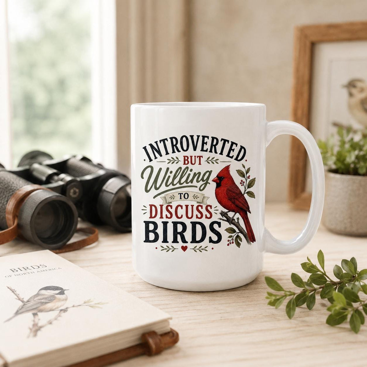 Cardinal mug on a wooden table with binoculars, a field guide showing a chickadee, a green sprig and a bird print