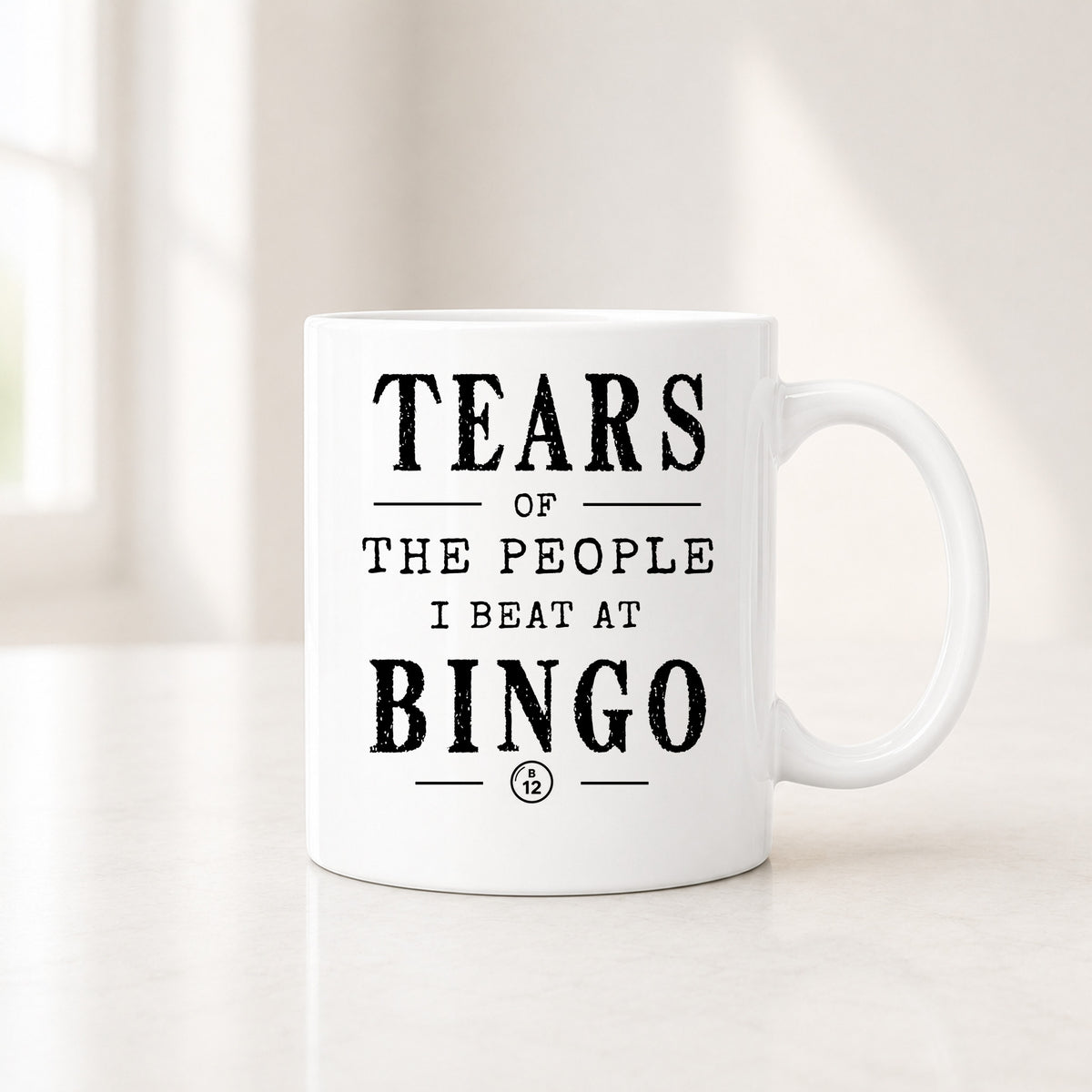 11 oz white bingo mug with the black Tears of the People I Beat quote, on a light counter by a window