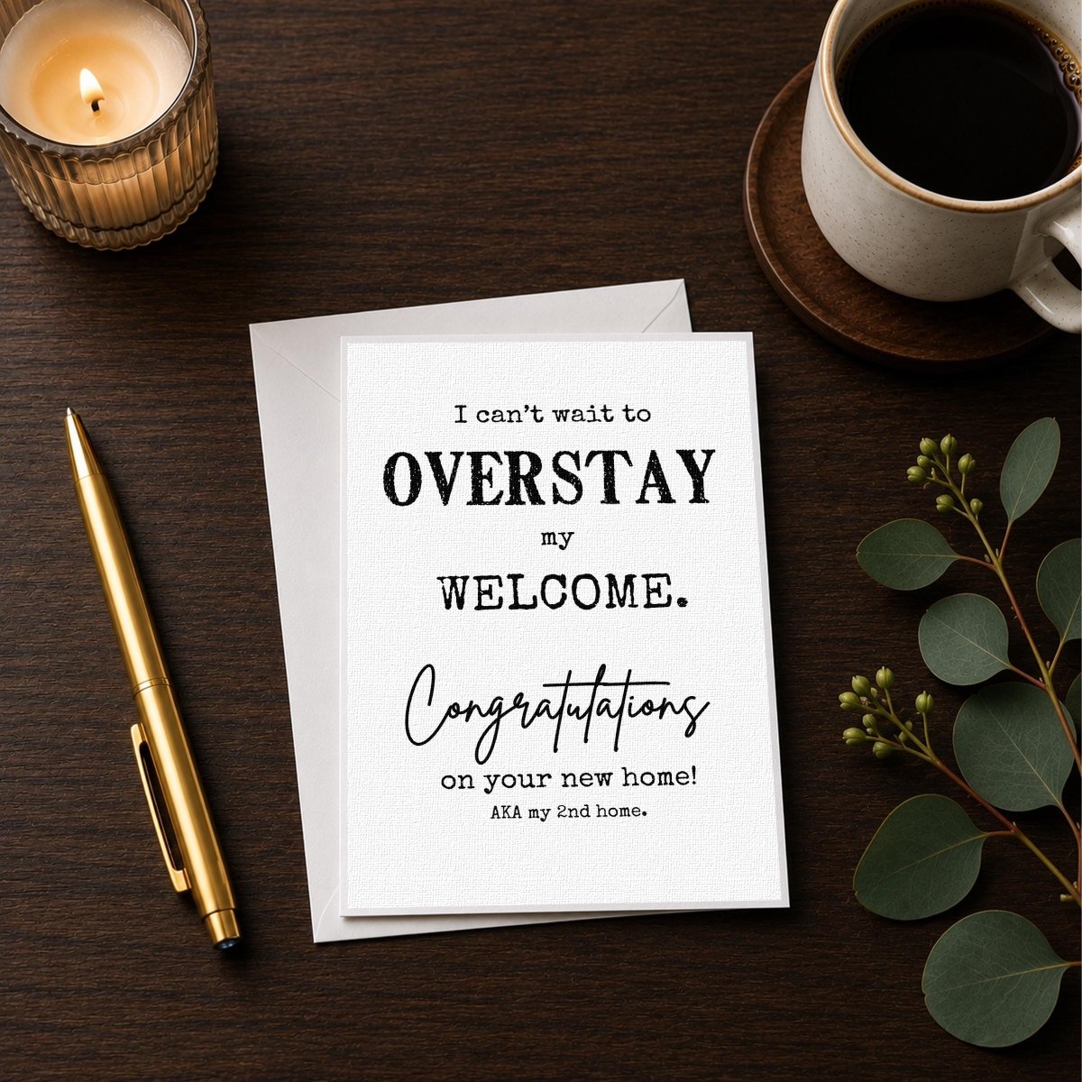 a greeting card with a message that reads "I can't wait to OVERSTAY my WELCOME. Congratulations on your new home! Aka my End home." The card is placed on a wooden surface alongside a cup of coffee and a pen.
