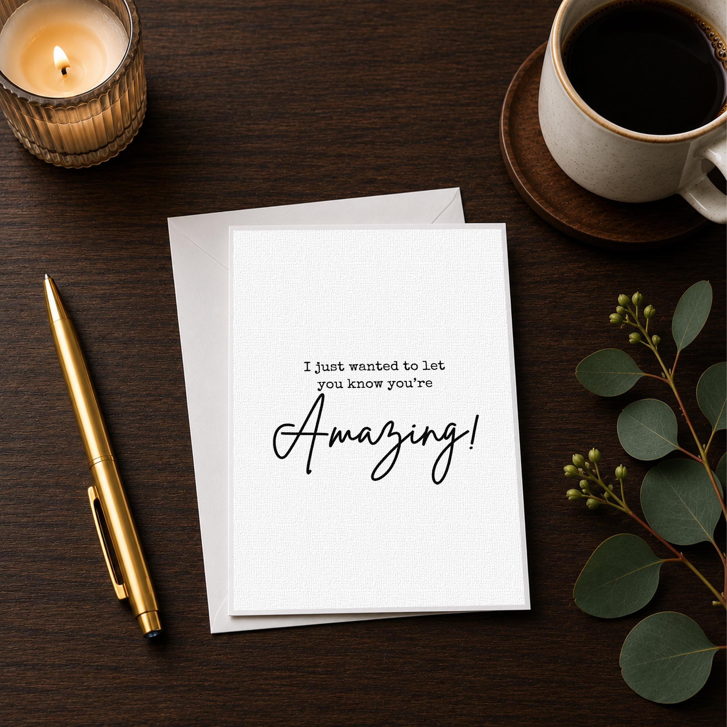 a white card with a message that says "I just wanted to let you know you're Amazing!" placed on a wooden surface alongside a candle, a pen, and a cup of coffee.