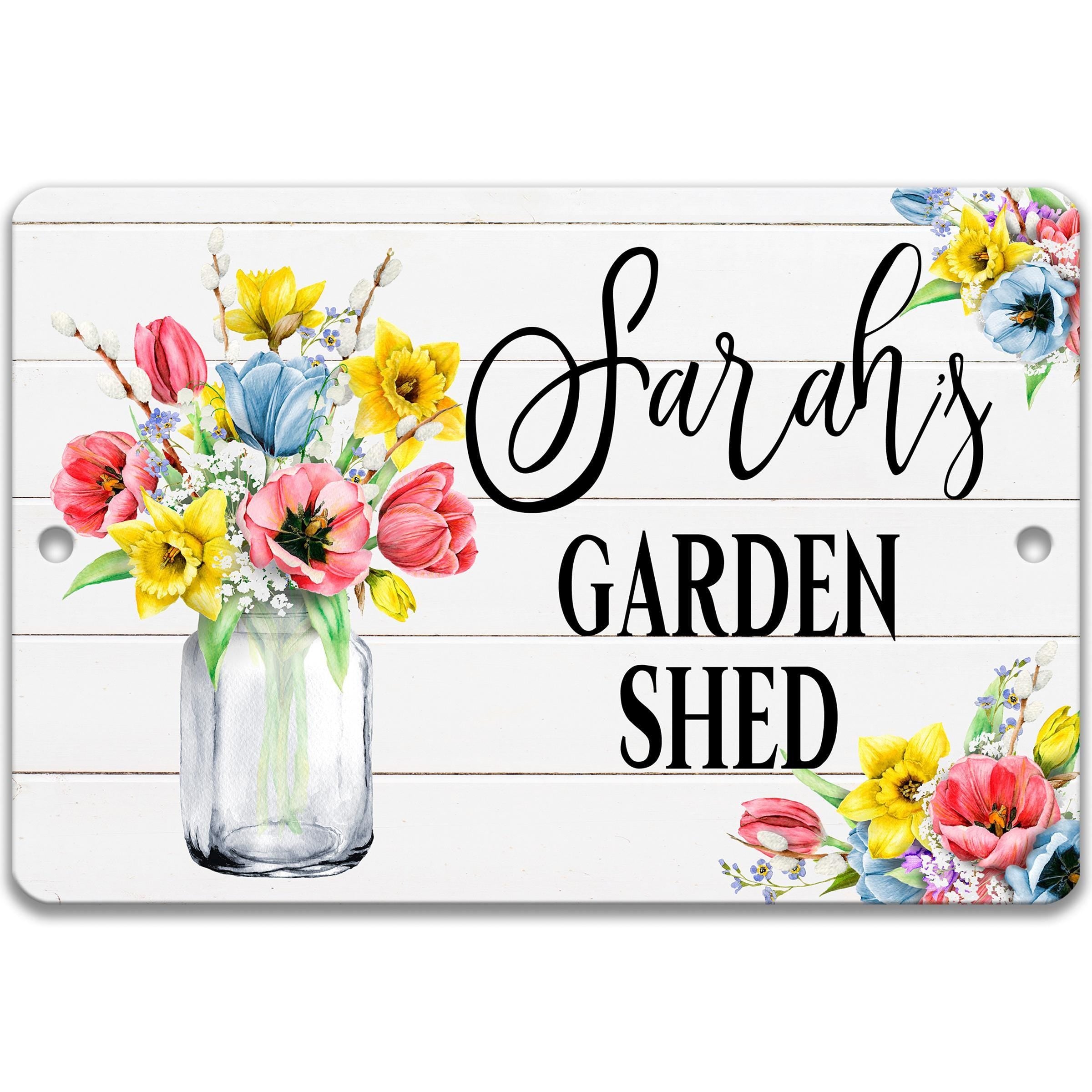 A white wooden sign with a vase of colorful flowers and the text "Sarah's GARDEN SHED" written on it.