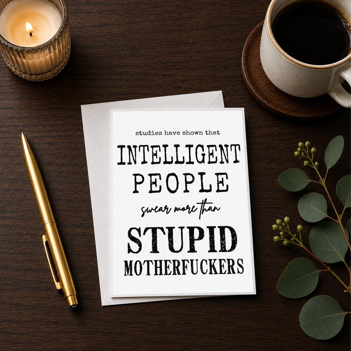 a card with a humorous message, a candle, a pen, and a cup of coffee on a wooden surface.