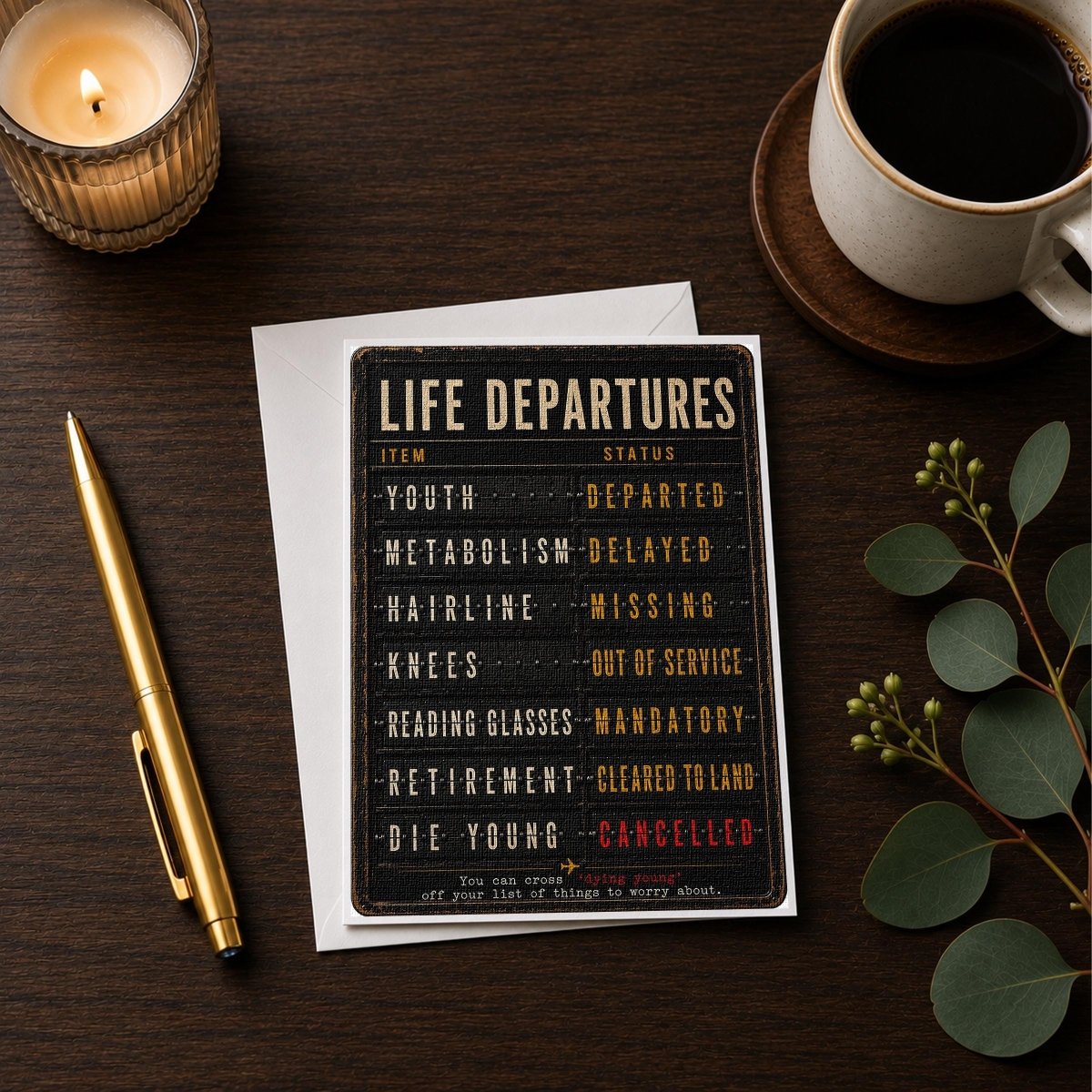 a card titled "Life Departures" with a list of various departures, such as youth, metabolism, hairline, knees, reading glasses, retirement, and dying young, along with a note about cancellations. The card is placed on a wooden surface, accompanied by a pen, a candle, and some greenery.