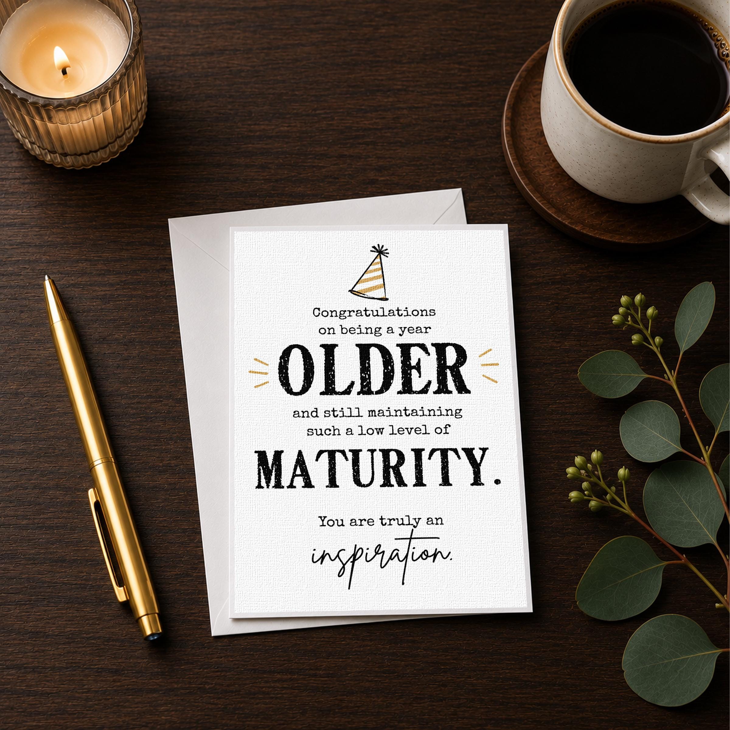 a card with a message congratulating an older person on being a year older and still maintaining a low level of maturity. The card is placed on a wooden surface next to a cup of coffee and a pen.