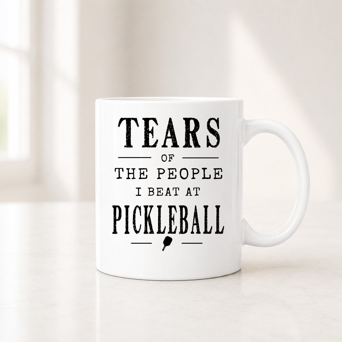 11 oz white pickleball mug with the black Tears of the People I Beat quote, on a light counter by a window
