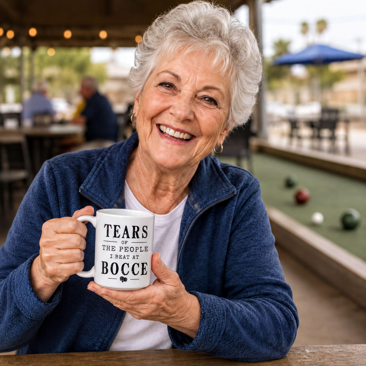 Smiling woman with gray hair holding the white Tears of the People I Beat at Bocce coffee mug
