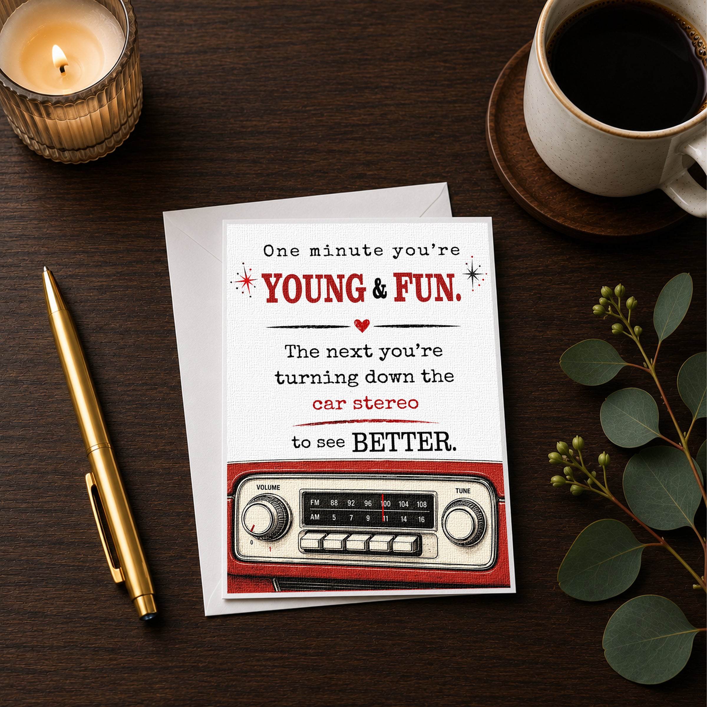 a card with a humorous message, a pen, a candle, a cup of coffee, and a radio on a wooden surface.