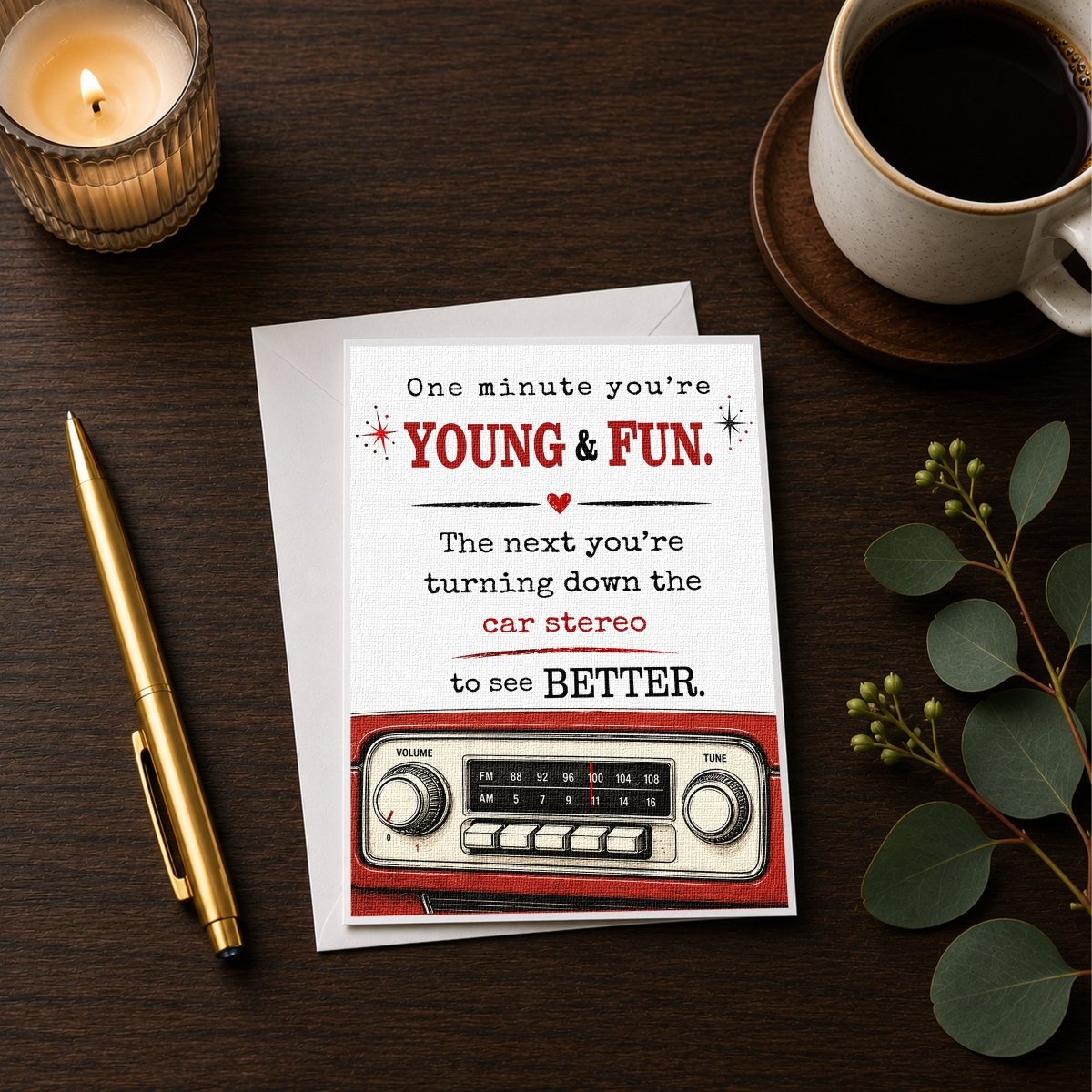a card with a humorous message, a pen, a candle, a cup of coffee, and a radio on a wooden surface.