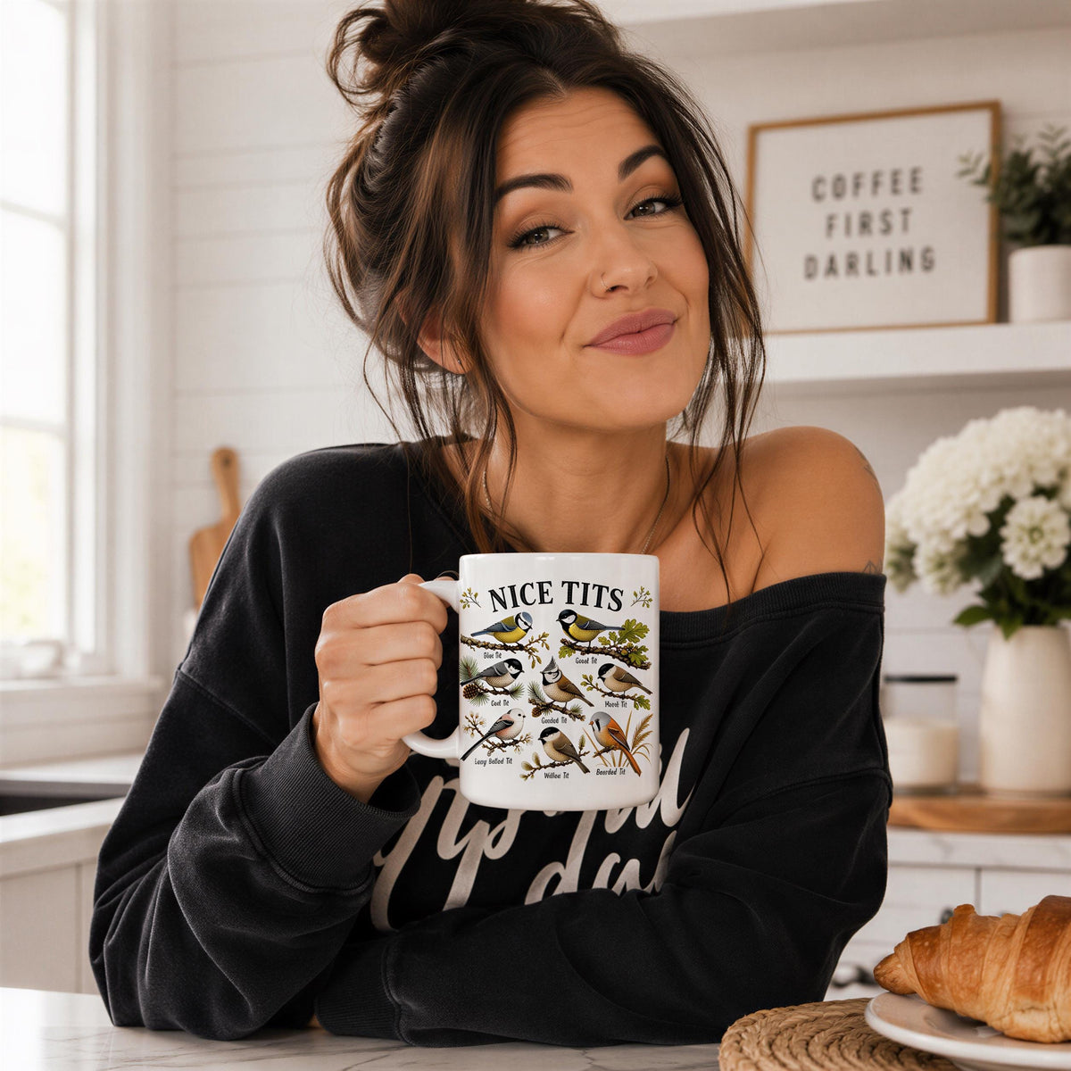 Woman with her hair in a bun smiling over the Nice Tits bird mug at a white kitchen counter beside a croissant