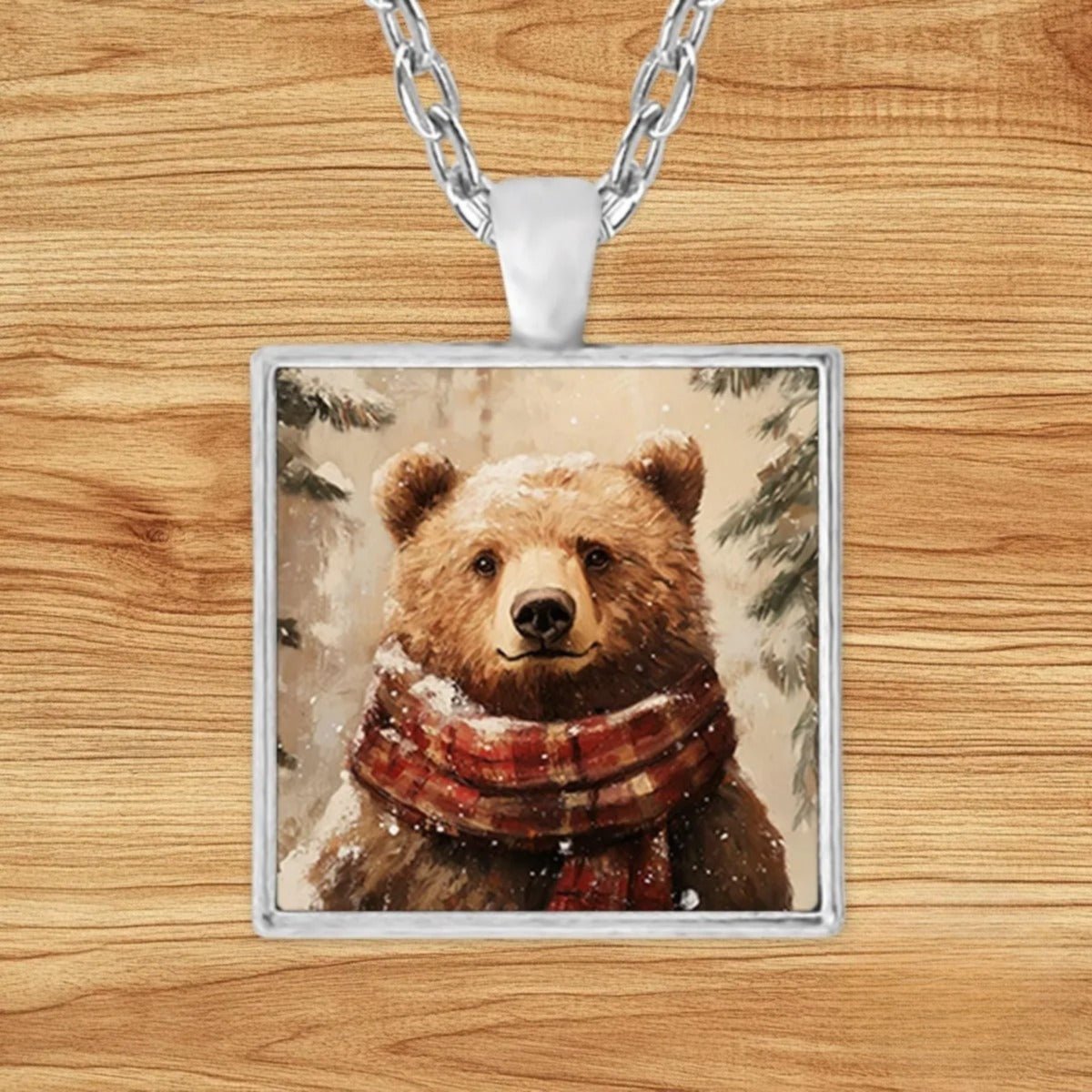 Square silver charm pendant necklace featuring a brown bear bundled in a red and white plaid scarf among snowy evergreens