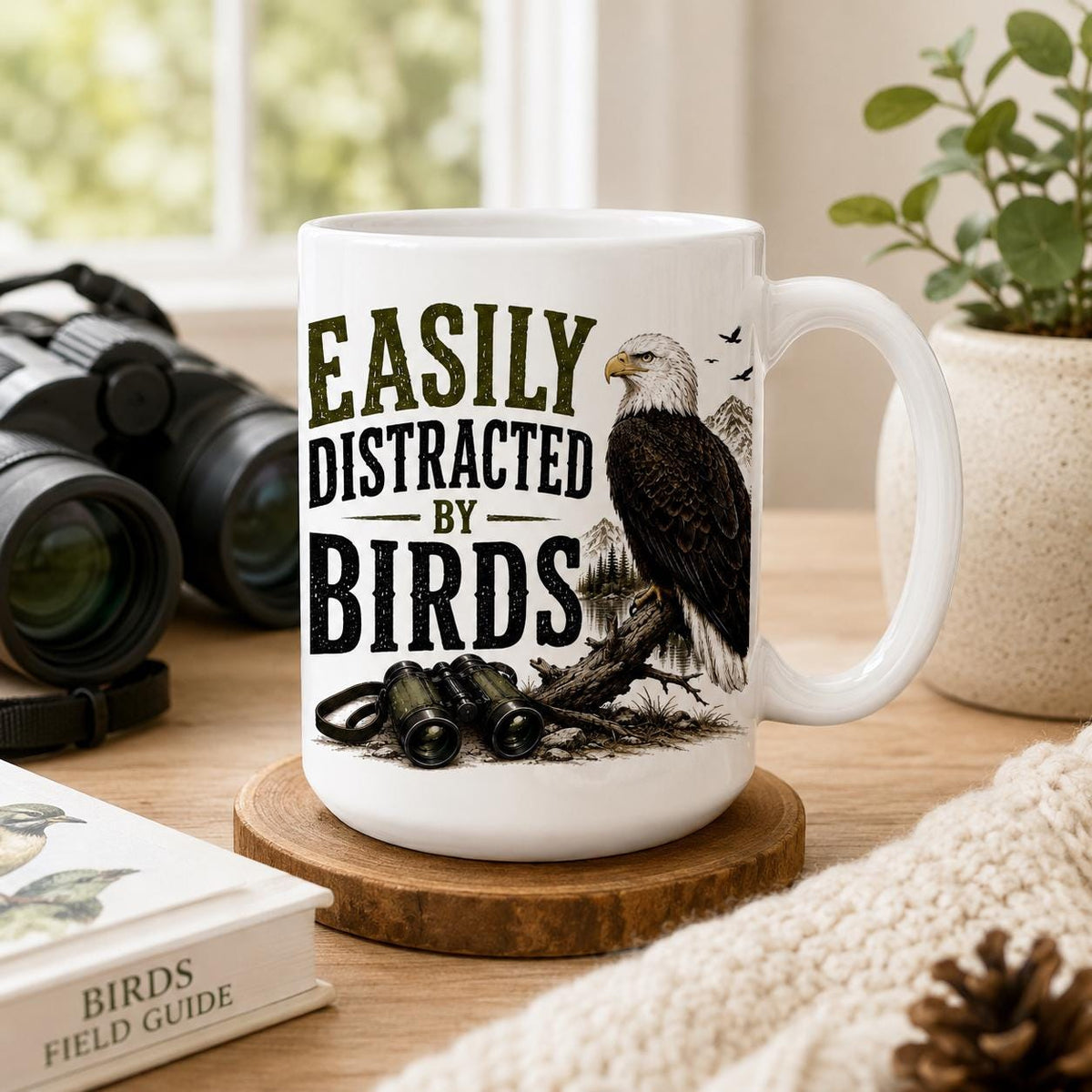 Easily Distracted by Birds mug on a wood coaster with binoculars, a birds field guide, a plant and a pinecone