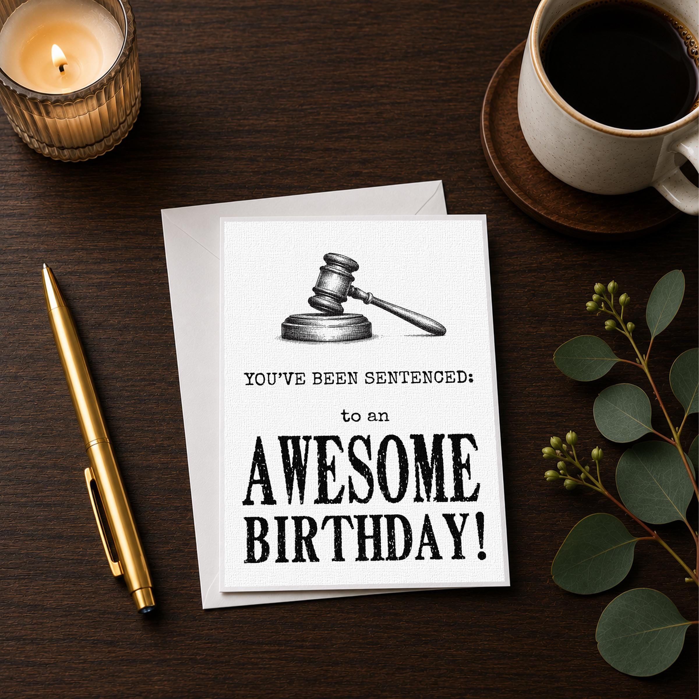 a birthday card with a gavel and the text "You've Been Sentenced: to an Awesome Birthday!" along with a pen and a candle on a wooden surface.