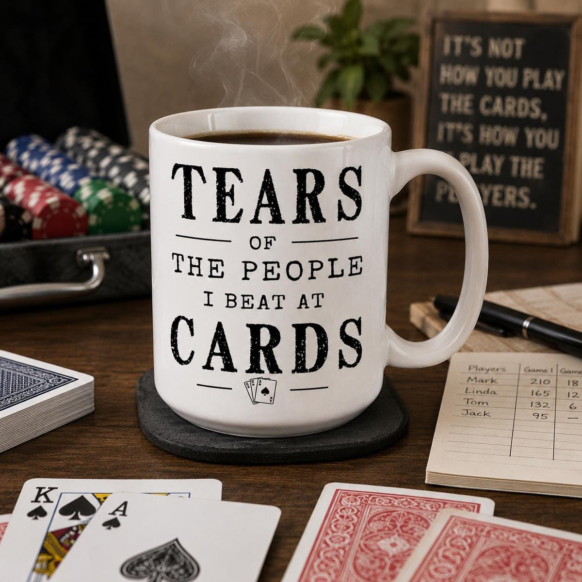 Card game mug on a wooden table surrounded by playing cards, a pen and a small plant
