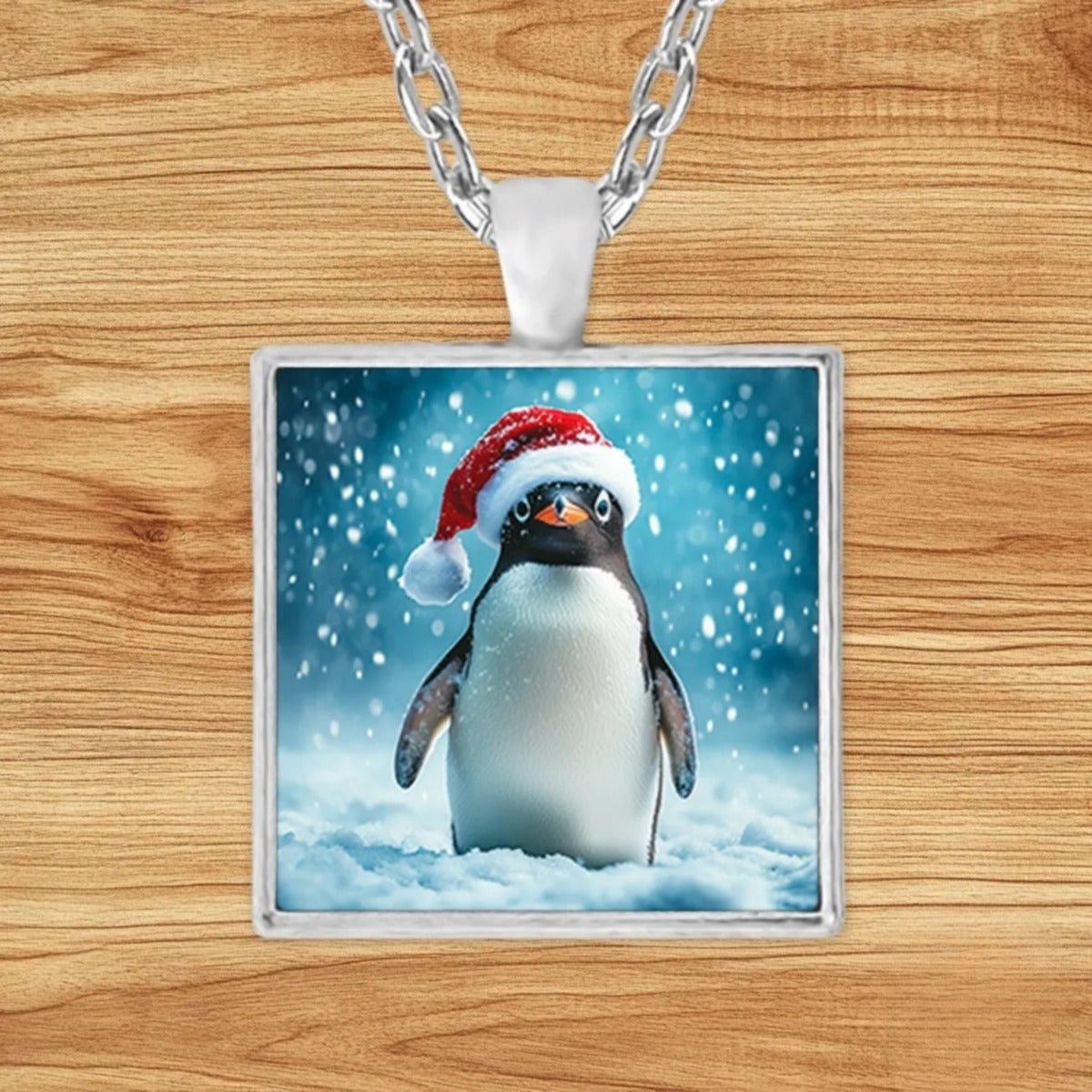 Square silver charm pendant necklace featuring a cheerful penguin in a bright red Santa hat amid swirling snowflakes
