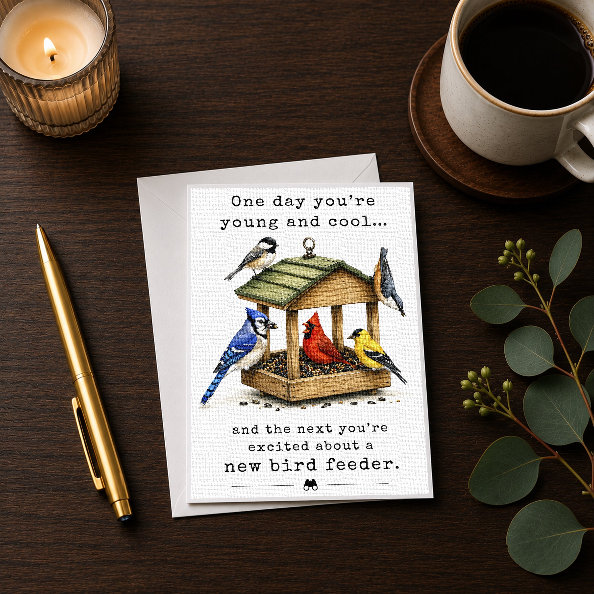 a greeting card with a birdhouse illustration, a pen, a candle, and a cup of coffee on a wooden surface.