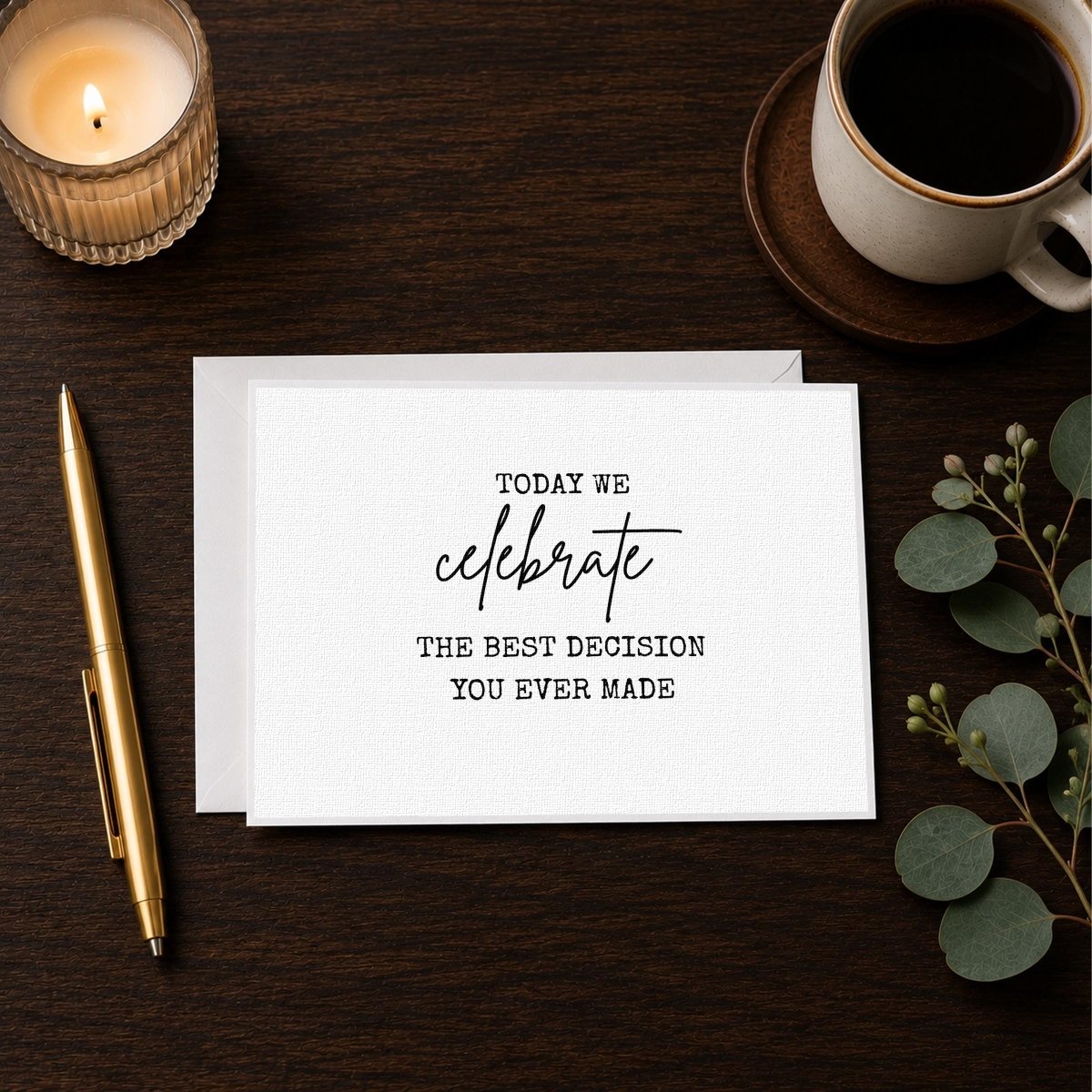 a white card with black text that reads "Today we celebrate, the best decision you ever made" placed on a wooden surface alongside a candle, a pen, and a cup of coffee.