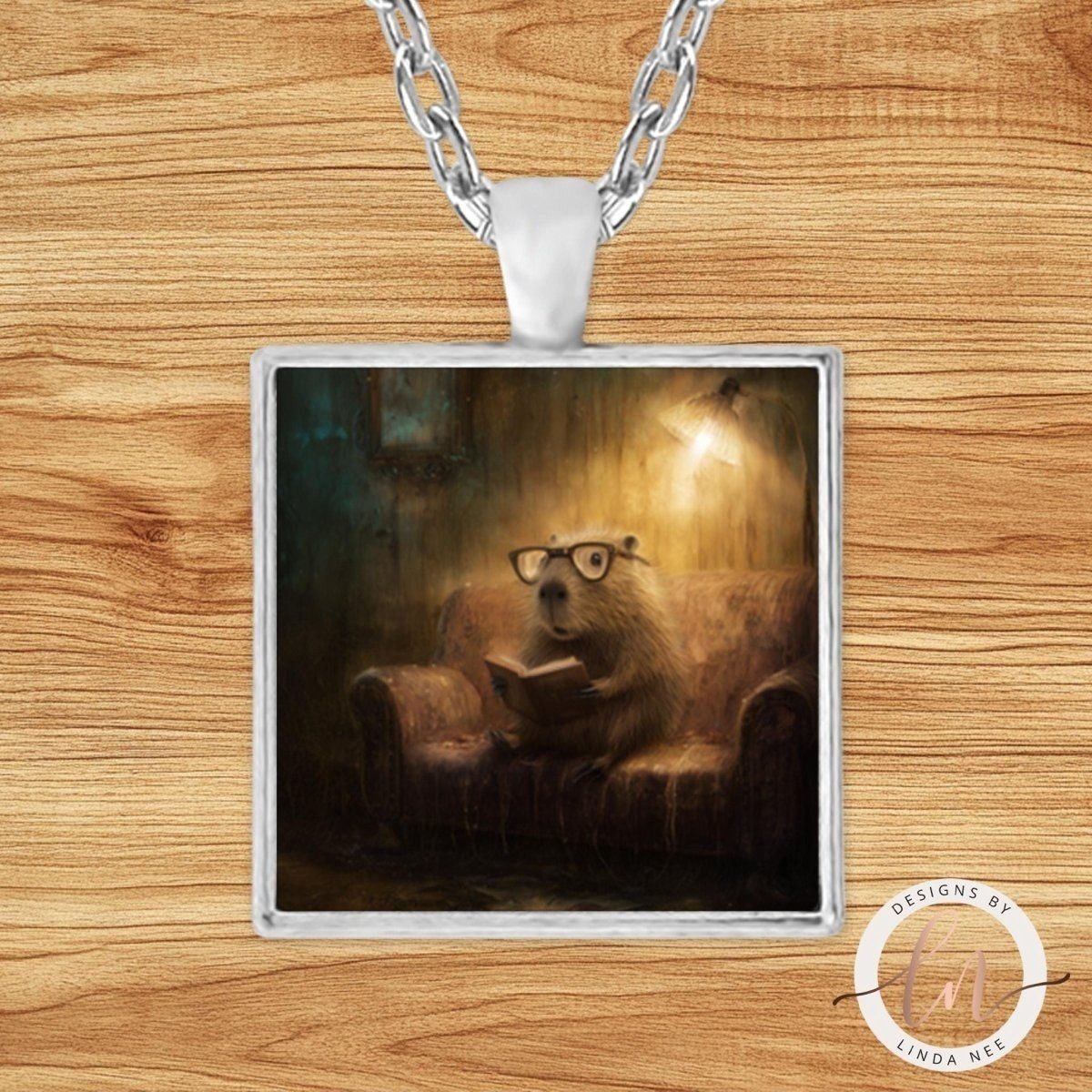 A pendant necklace with a square frame containing an illustration of a beaver wearing glasses and reading a book, set against a wooden background.