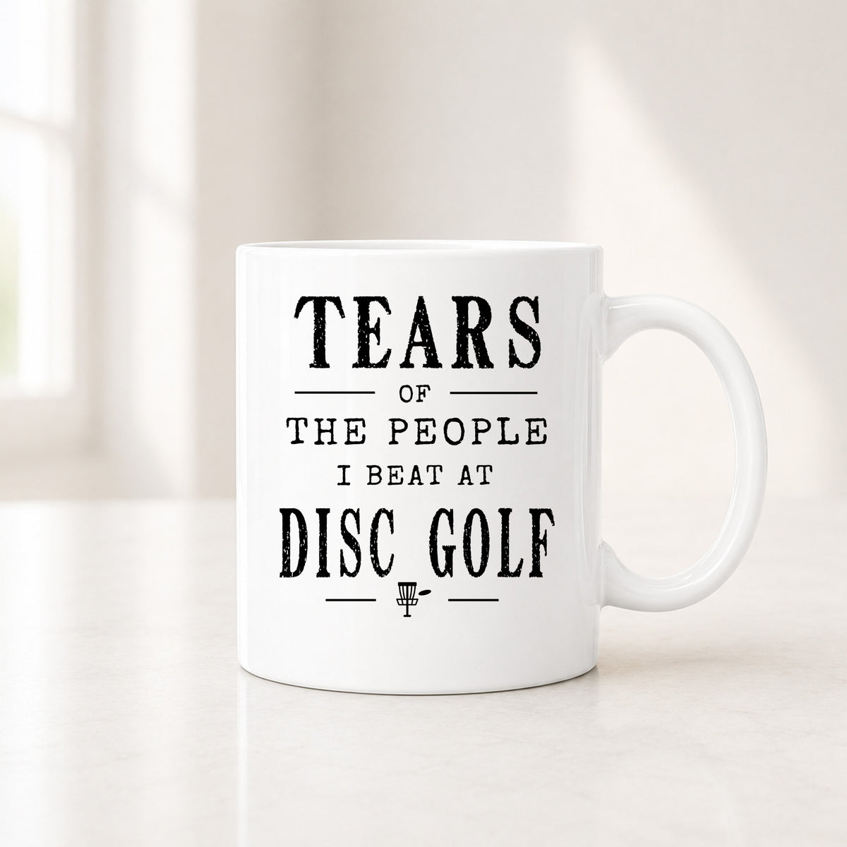 11 oz white disc golf mug with the black Tears of the People I Beat quote, on a light counter by a window