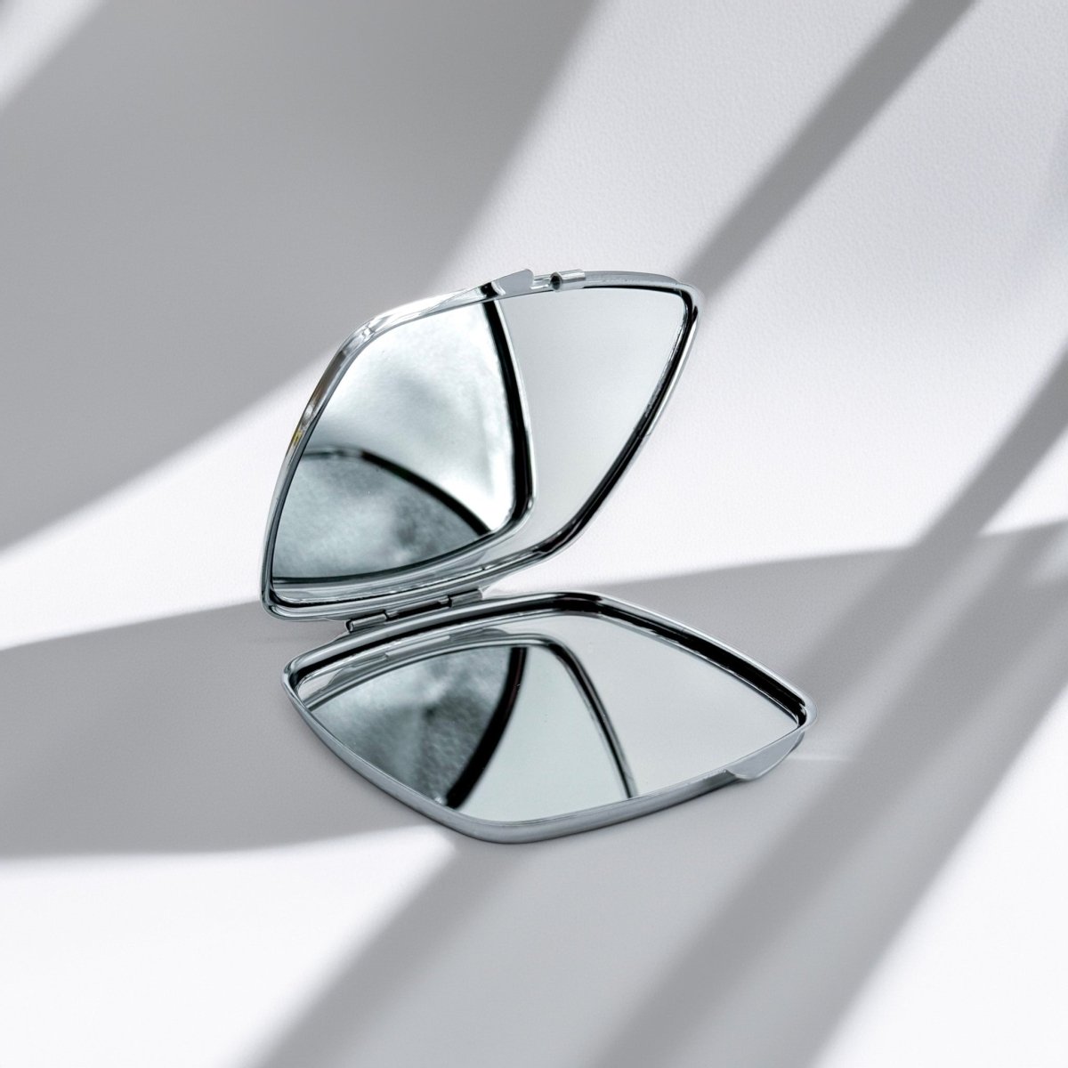 A compact mirror with a silver frame is open and resting on a white surface, reflecting the light.