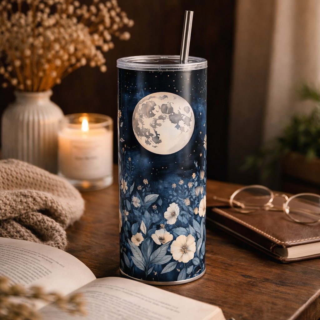 A tall, cylindrical tumbler with a floral design and a silver straw sits on a wooden surface, surrounded by various objects such as a candle, glasses, and a book.