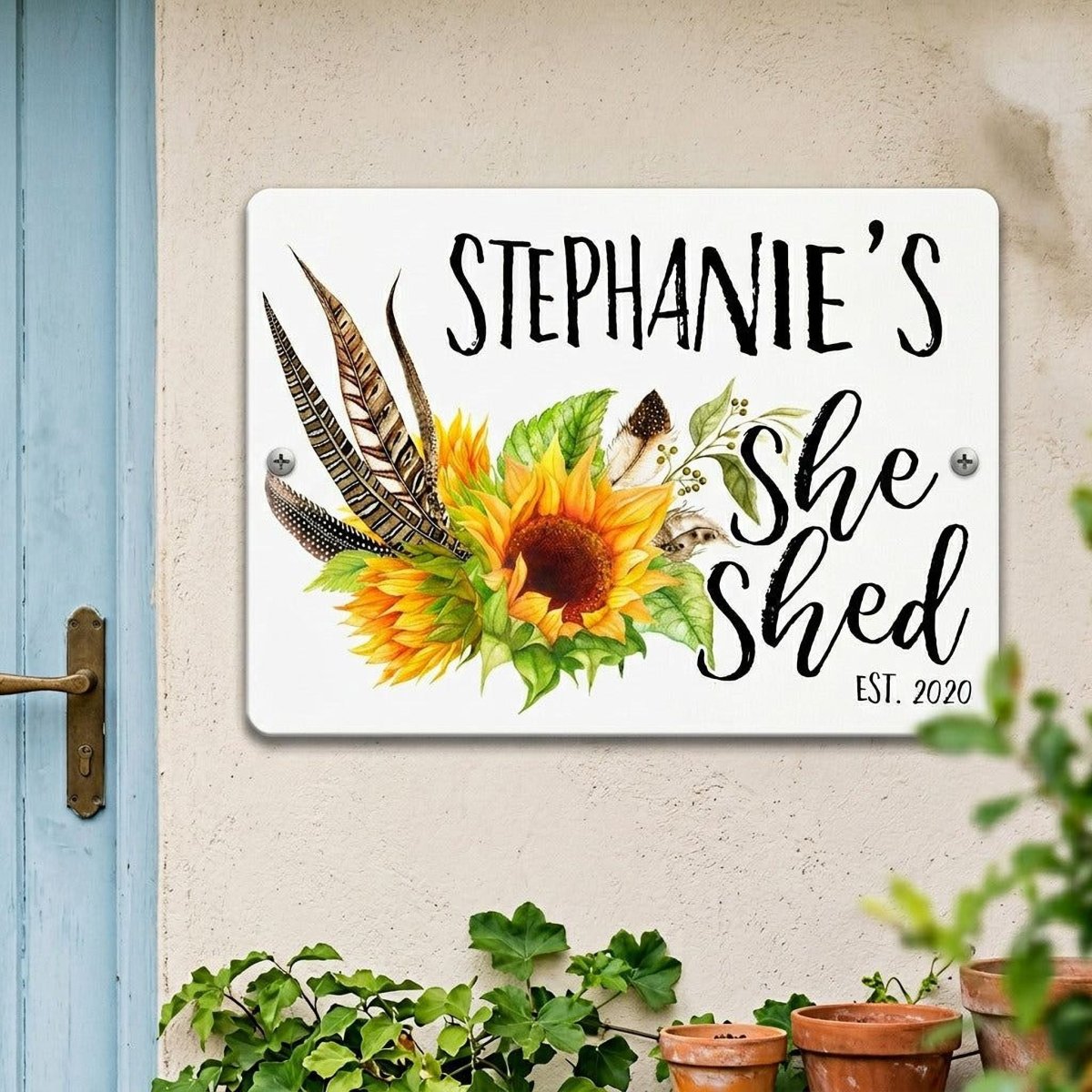 A white sign with a sunflower and feathers hanging on a wall, with the text "Stephanie's She Shed" and "EST. 2020" below it.