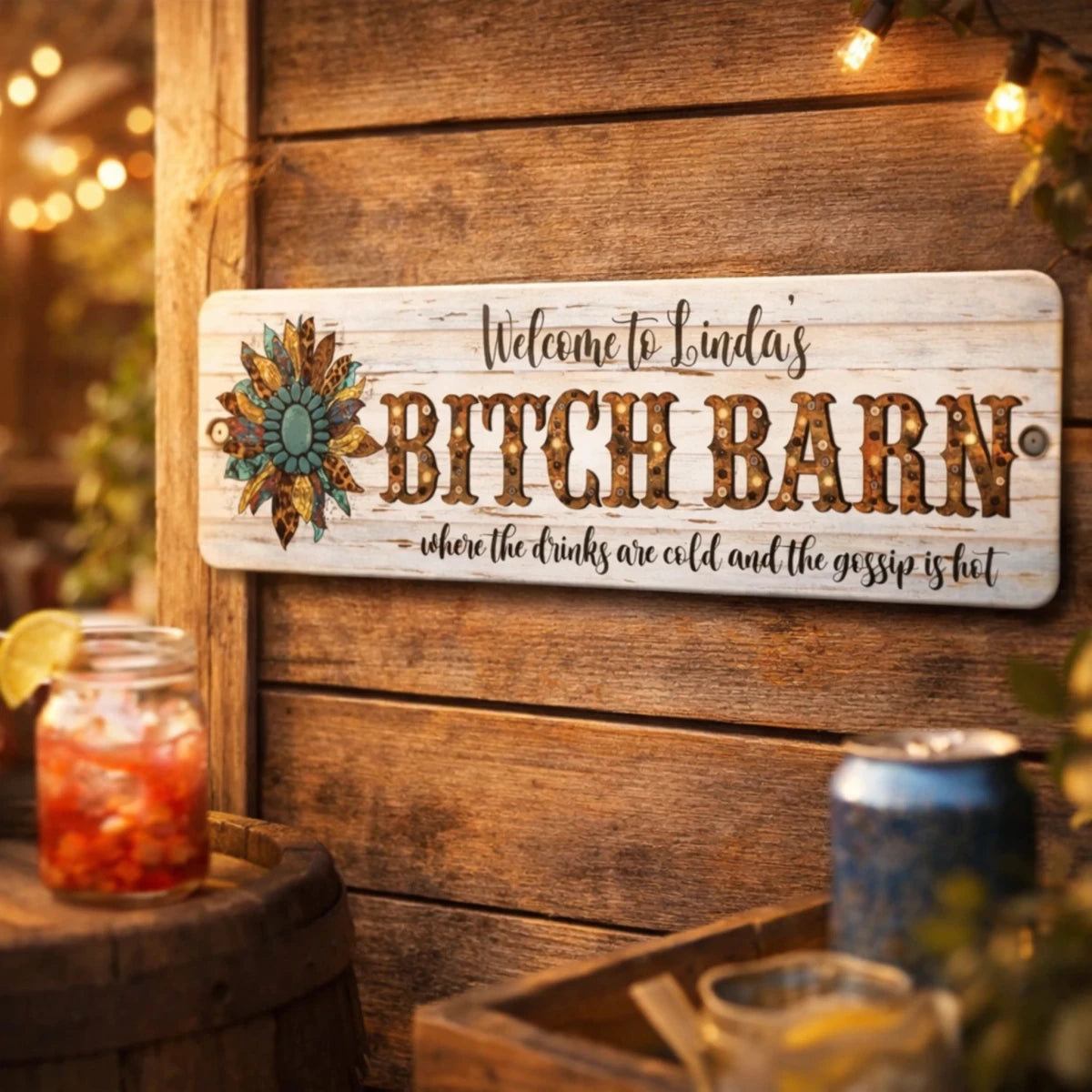 a wooden sign with a decorative design, hanging on a wooden wall. The sign reads "Welcome to Linda's Bitch Barn" and includes a decorative element of a flower. In the background, there are some drinks, including a glass of red liquid and a glass of blue liquid, as well as a can of soda.