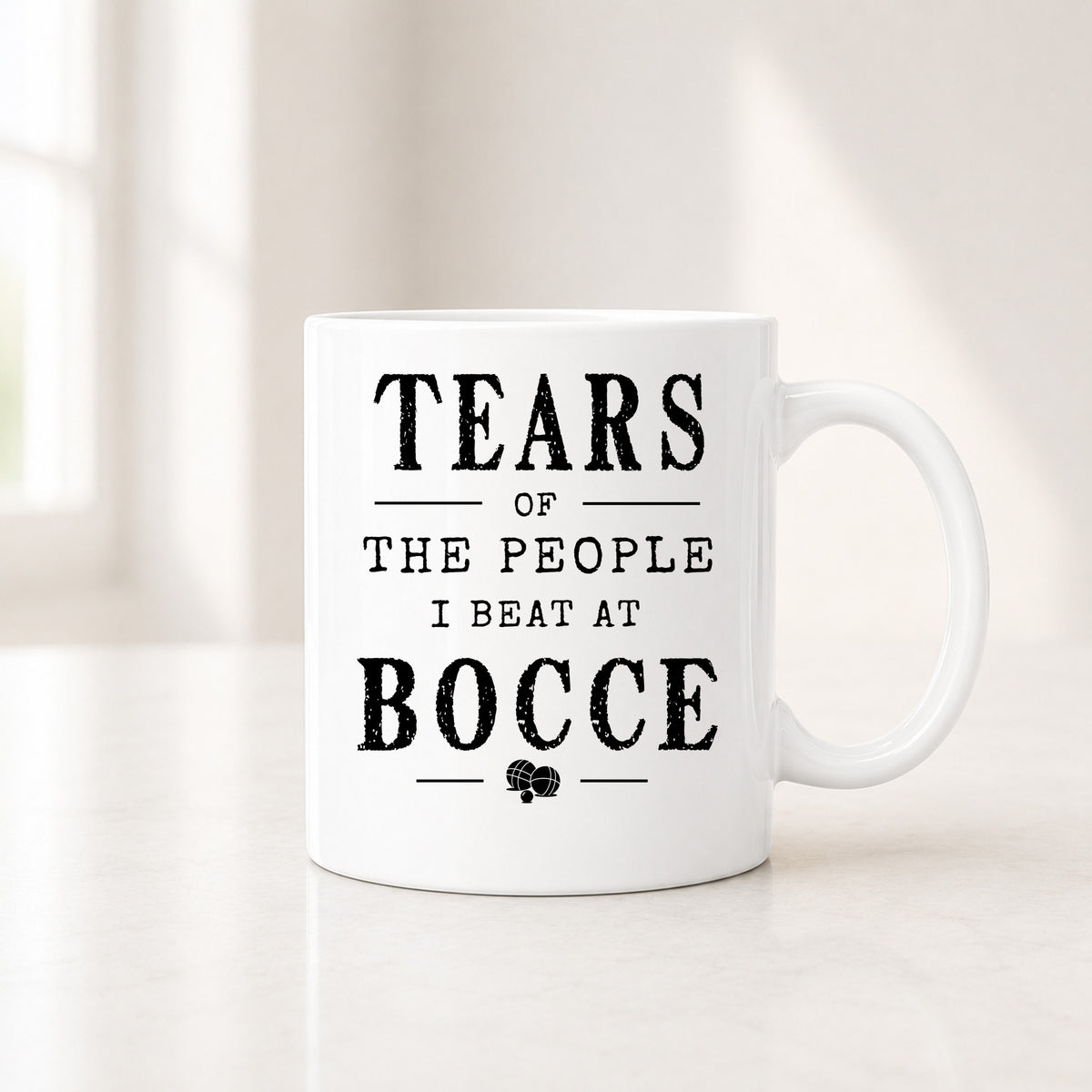 11 oz white bocce mug with the black Tears of the People I Beat quote, on a light counter by a window