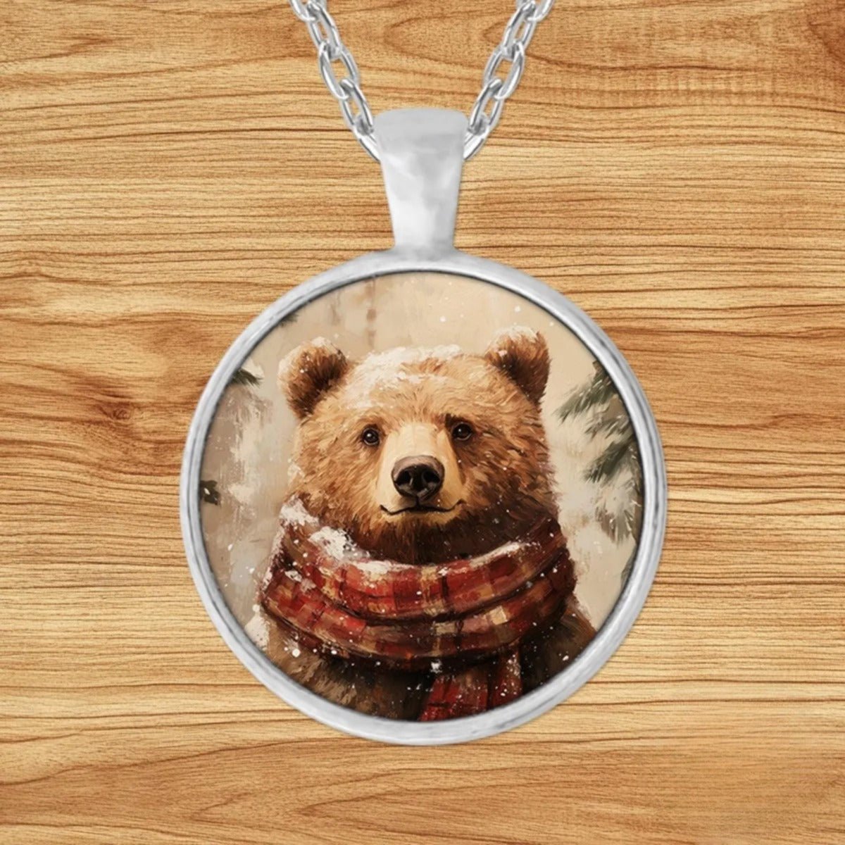 Round silver charm pendant necklace featuring a brown bear bundled in a red and white plaid scarf among snowy evergreens
