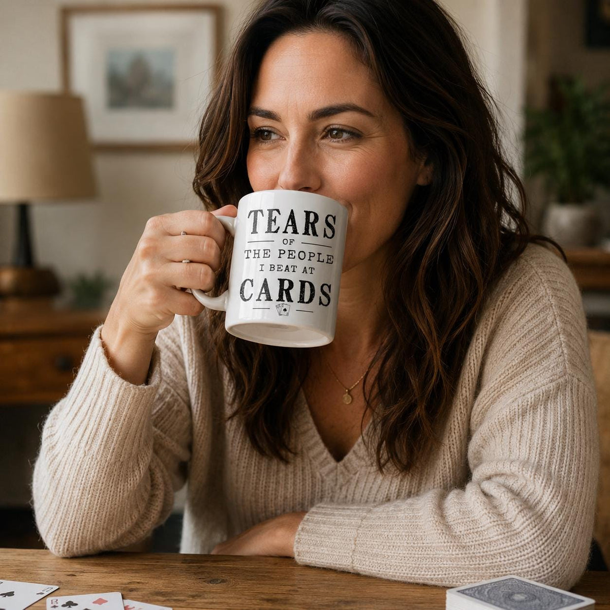 Woman sitting at a table holding the white Tears of the People I Beat at Cards coffee mug