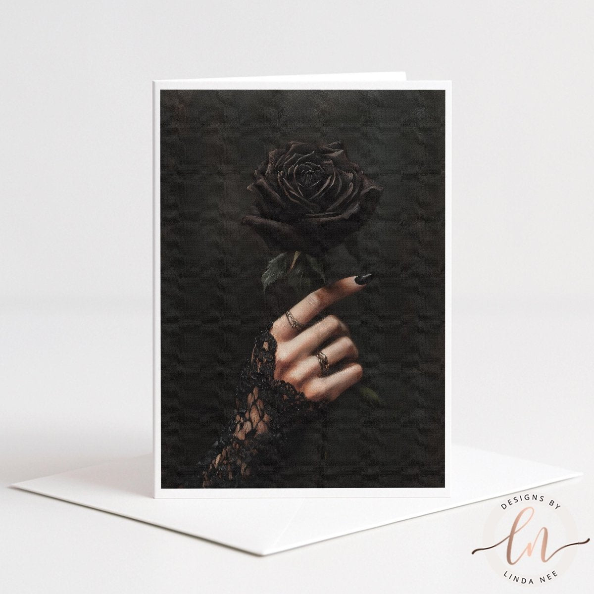 A vertical greeting card with a white border stands upright on a white envelope against a light gray background. The card features a dark, moody photograph of a hand with black nail polish and rings wearing a black lace glove, holding a single black rose.