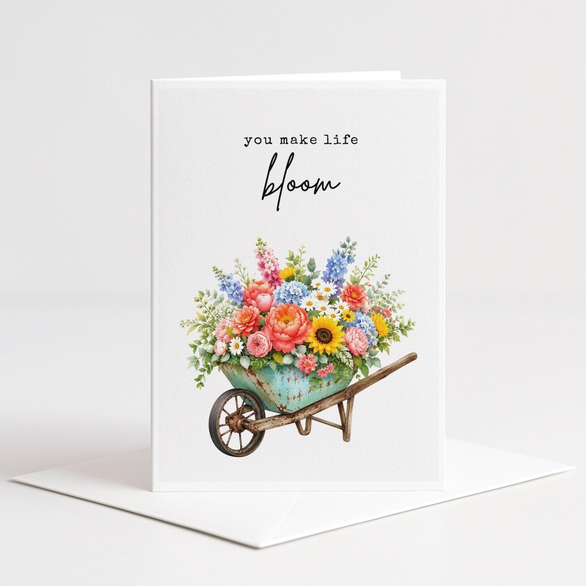 White 5-by-7 greeting card featuring the words “You make life bloom” above a vintage turquoise wheelbarrow overflowing with colorful garden flowers.