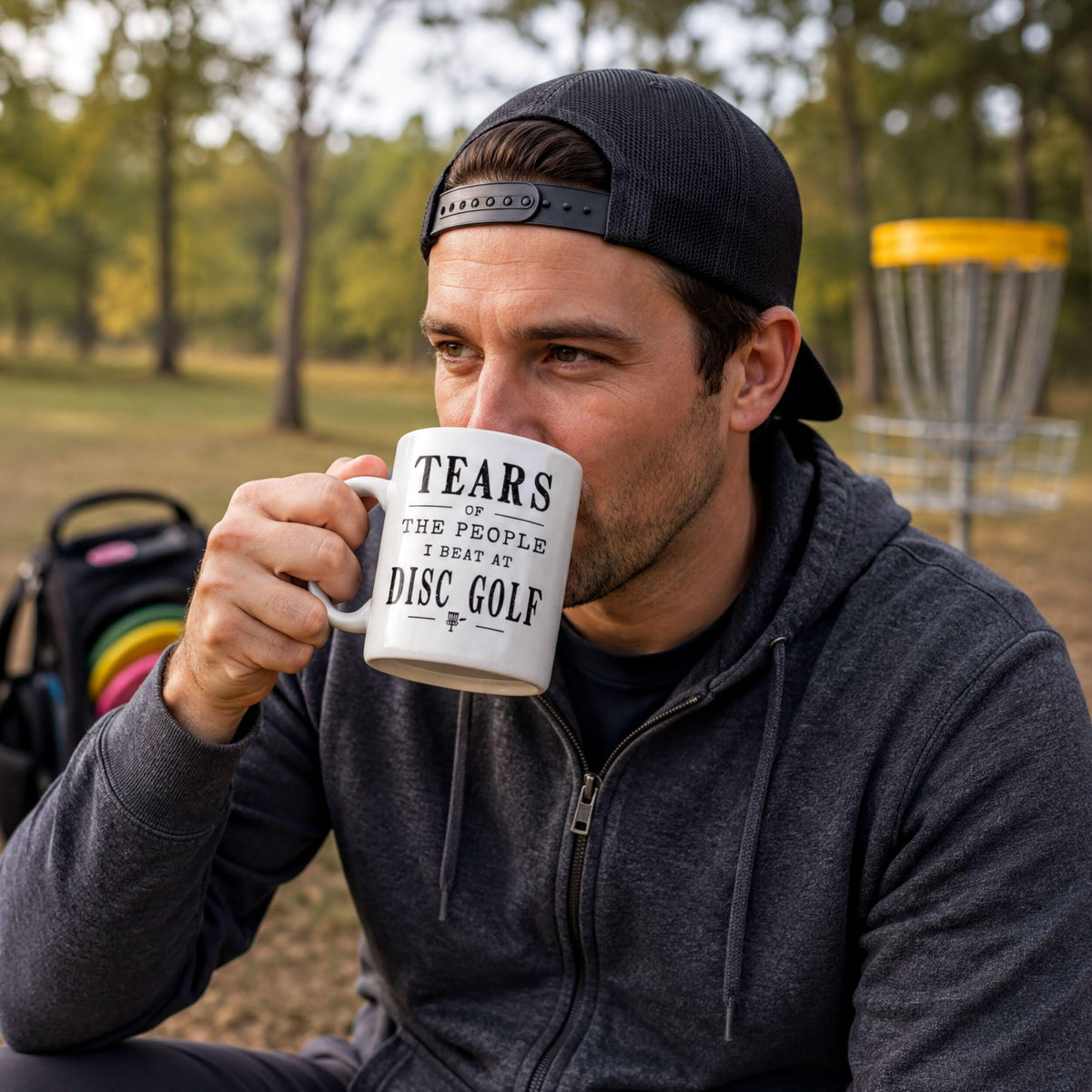 Man in a black cap sitting in a park and drinking from the Tears of the People I Beat at Disc Golf mug