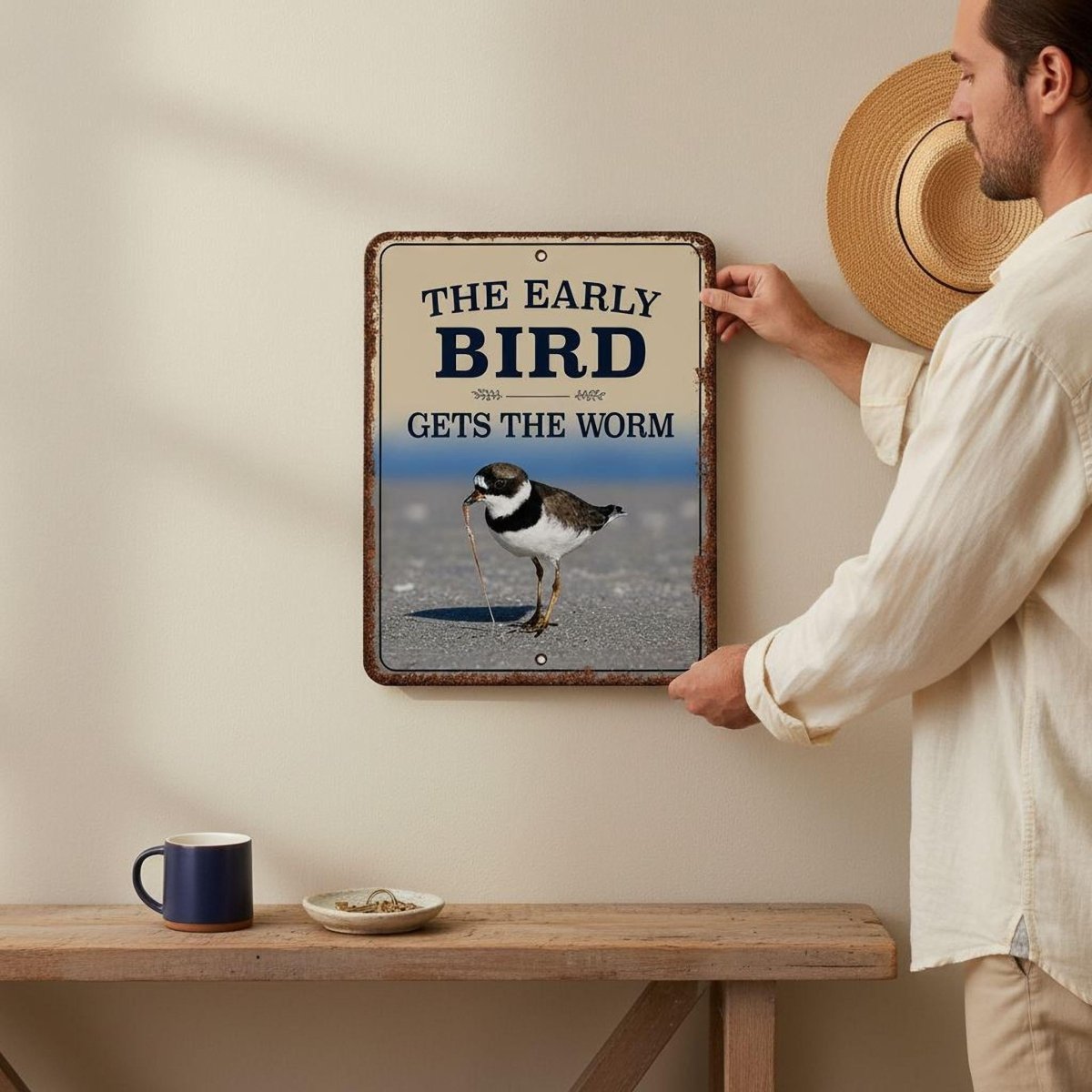 A man is hanging a metal sign on a wall, which features a bird and the text "The early bird gets the worm".