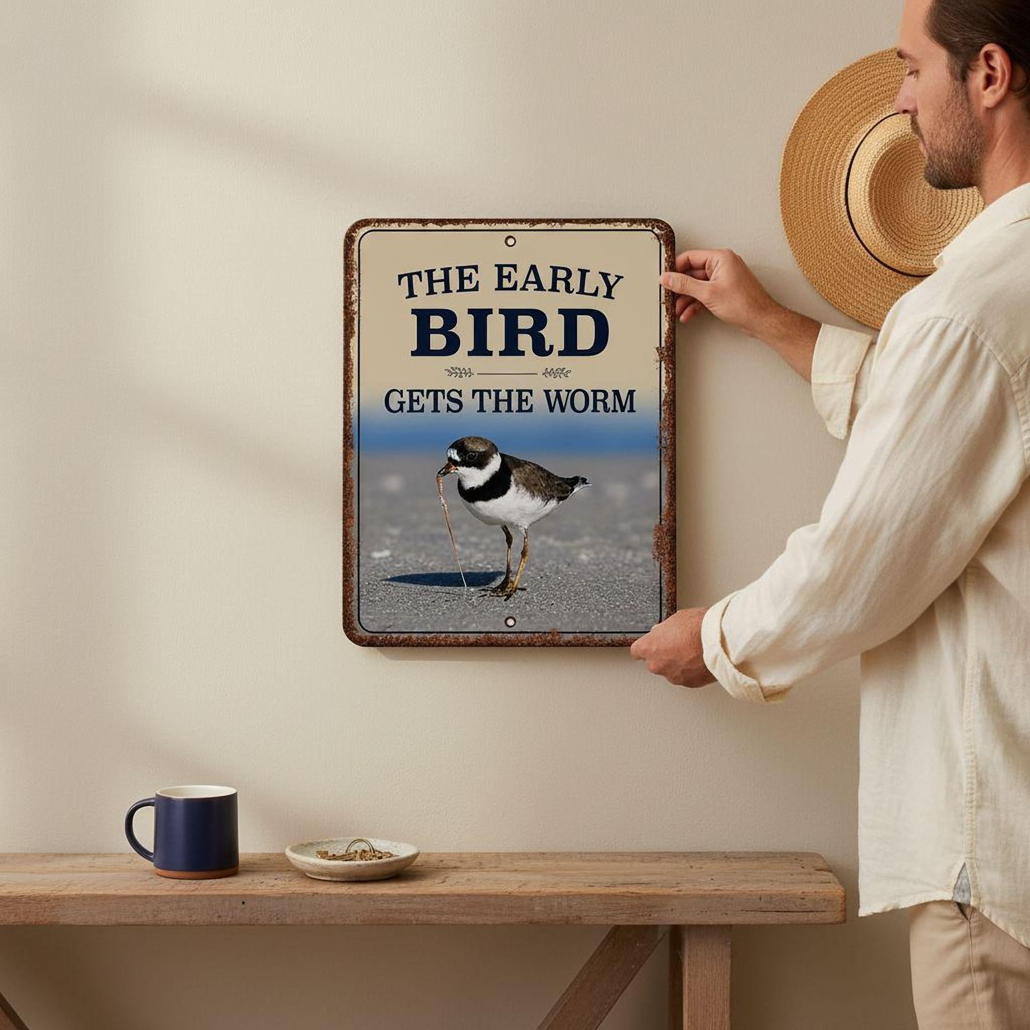 A man is hanging a metal sign on a wall, which features a bird and the text "The early bird gets the worm".