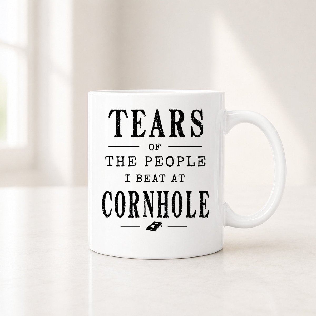 11 oz white cornhole mug with the black Tears of the People I Beat quote, on a light counter by a window