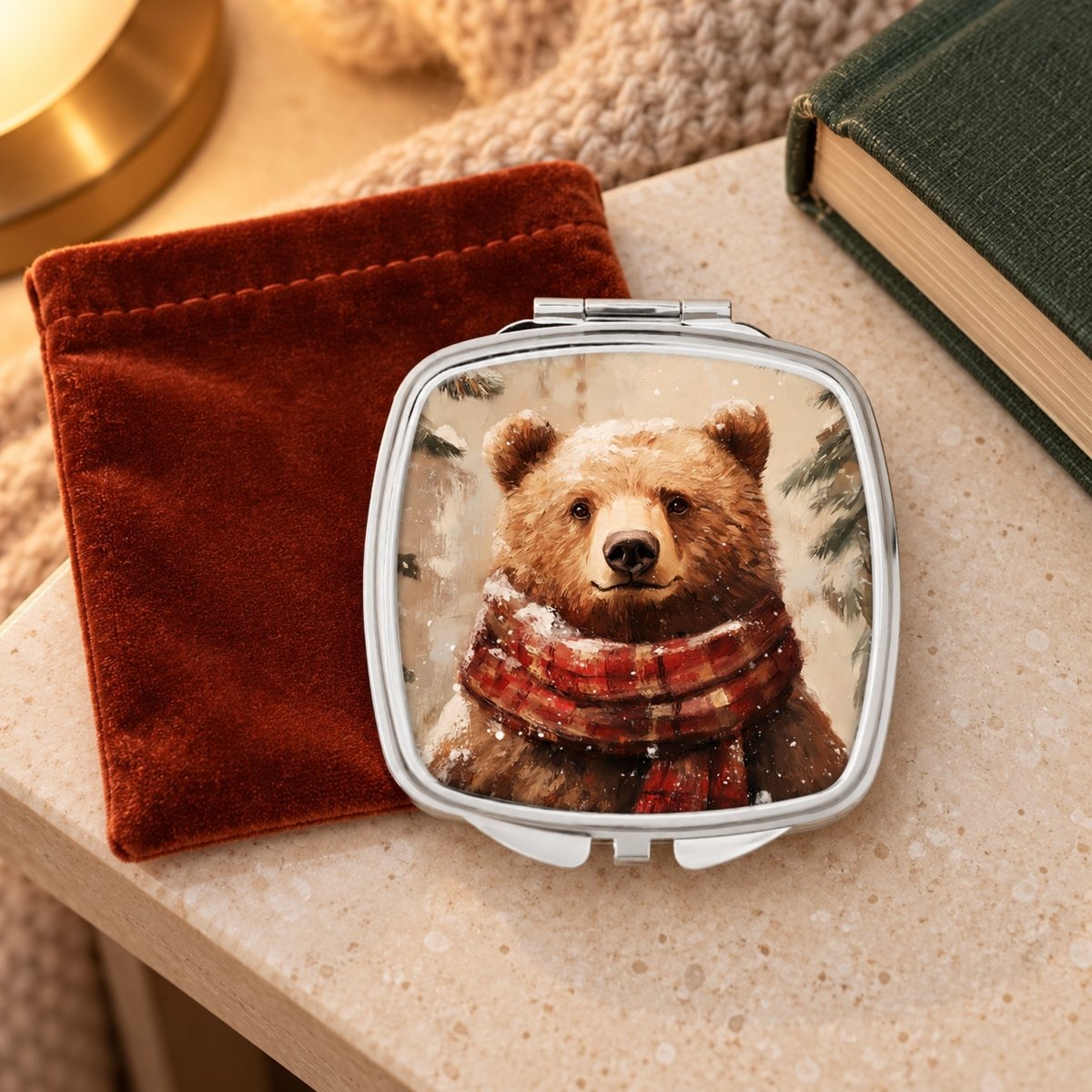 A compact mirror with a picture of a brown bear wearing a red scarf is placed on a table next to a red velvet pouch and a green book.