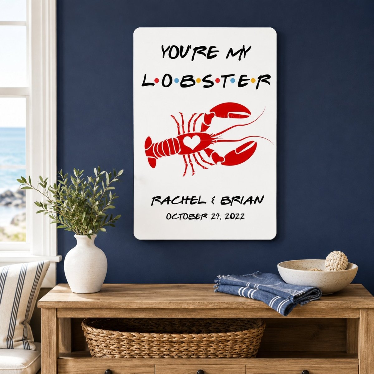 a wall with a framed poster that says "YOU'RE MY LOBSTER" and "RACHEL & BRIAN OCTOBER 24, 2023". Below the poster, there is a wooden table with a vase of green leaves, a bowl, and a striped towel.