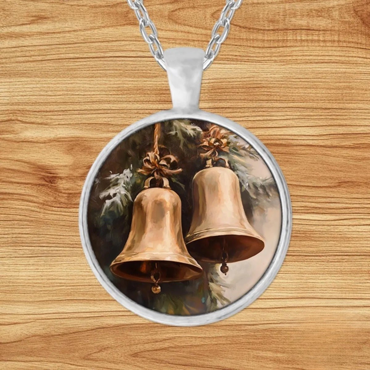Round silver charm pendant necklace featuring two brass bells tied with rope and bows hanging from snow-kissed pine branches