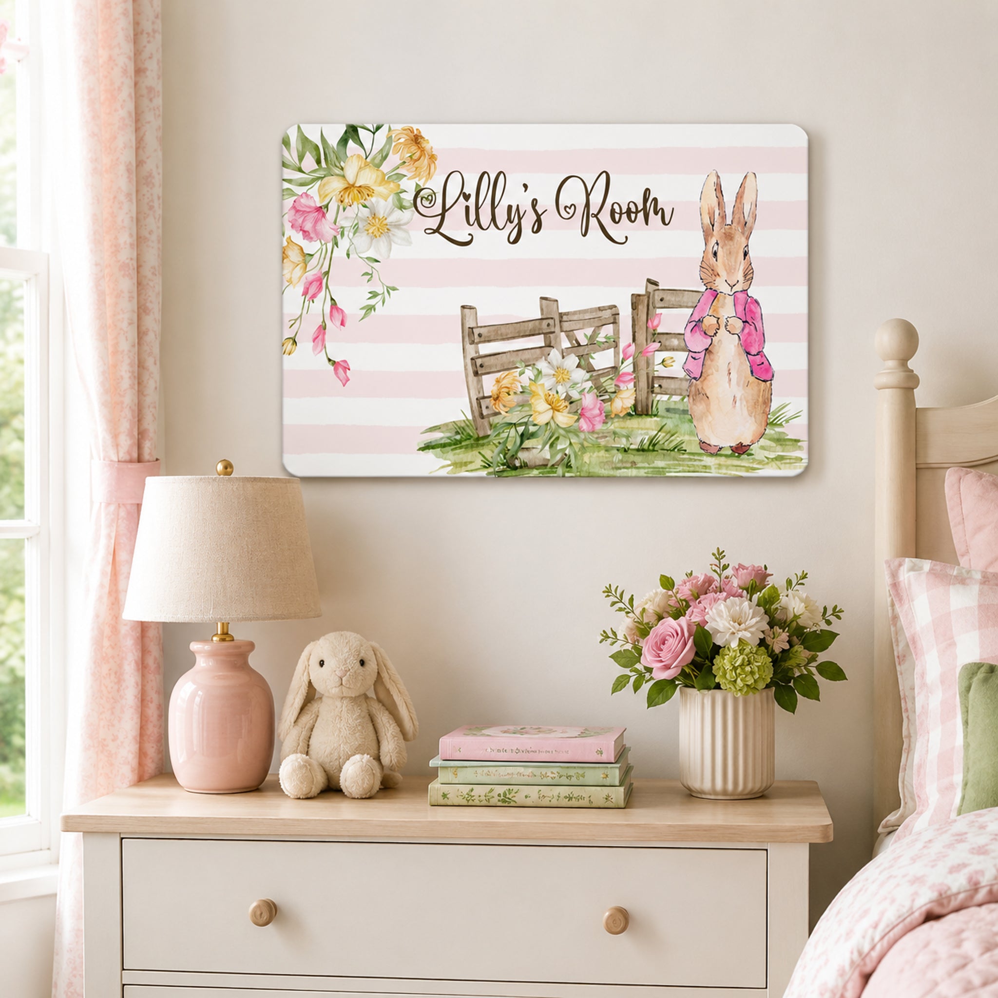 A bedroom with a pink and white color scheme, featuring a wooden dresser with a lamp, a stuffed rabbit, and a vase of flowers. Above the dresser is a framed print with a bunny and a fence, and the text "Lilly's Room" is written on the print.