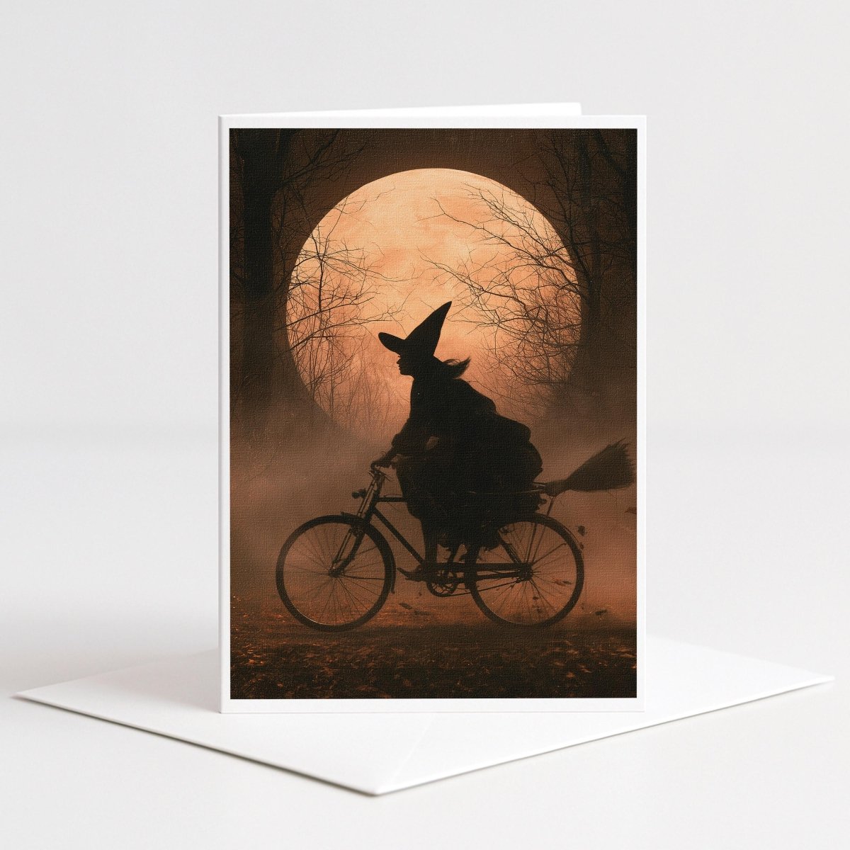 Witch silhouette riding a bicycle past a glowing orange moon in a misty autumn forest, note card art
