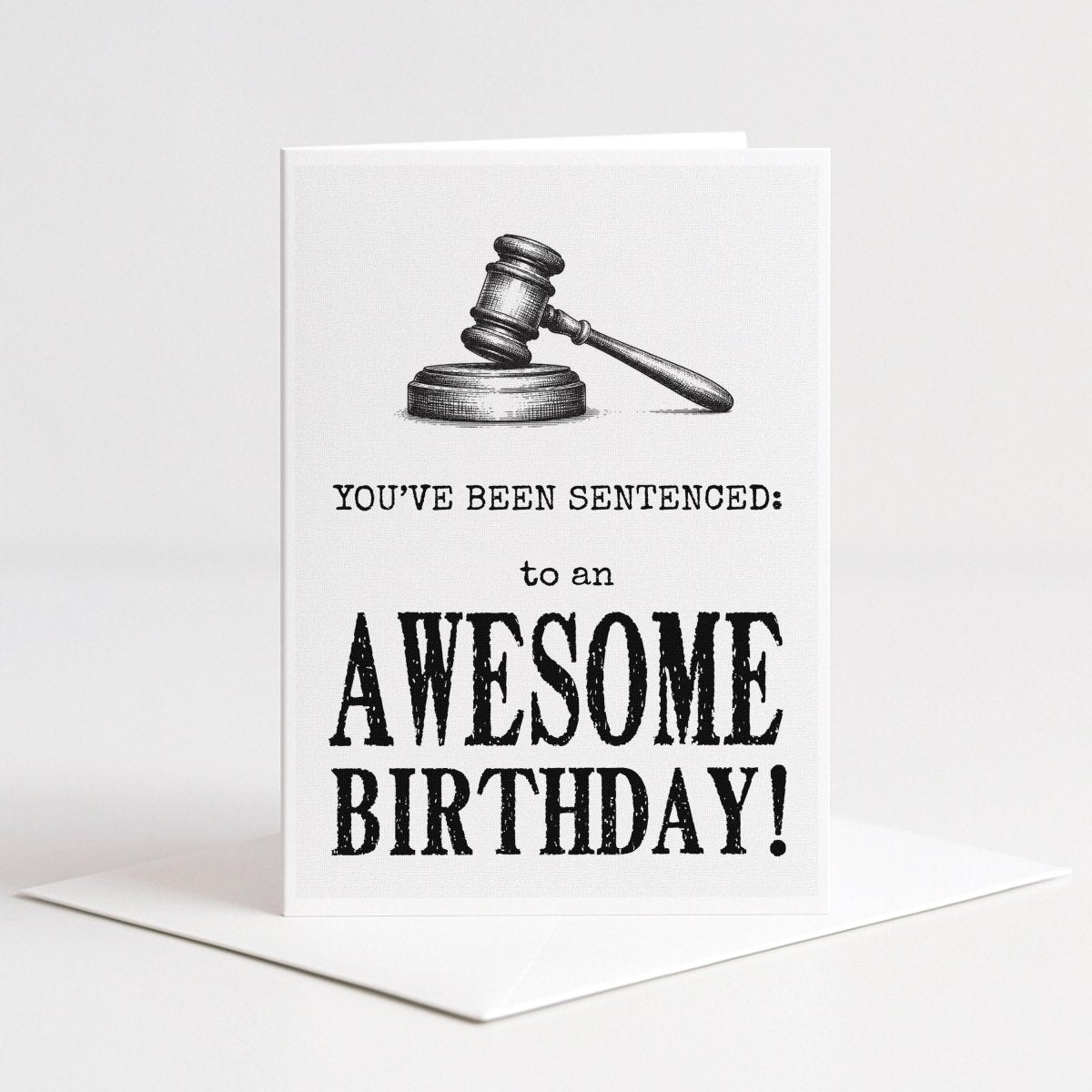 White 5-by-7 greeting card displayed upright on a white envelope with a detailed black gavel illustration and distressed typography reading, “You’ve been sentenced to an awesome birthday!”