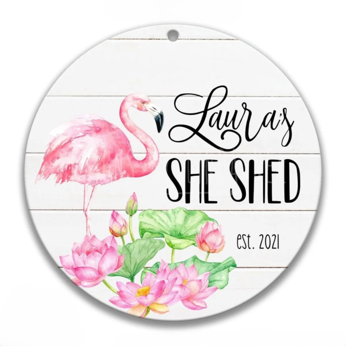 A round white sign with a pink flamingo and pink lotus flowers, along with the text "Lauren's She Shed est. 2021".