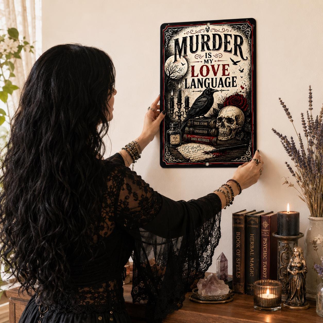 A woman with long dark hair is hanging a framed poster on a wall, which features a skull and a rose, along with the text "MURDER IS MY LOVE LANGUAGE".