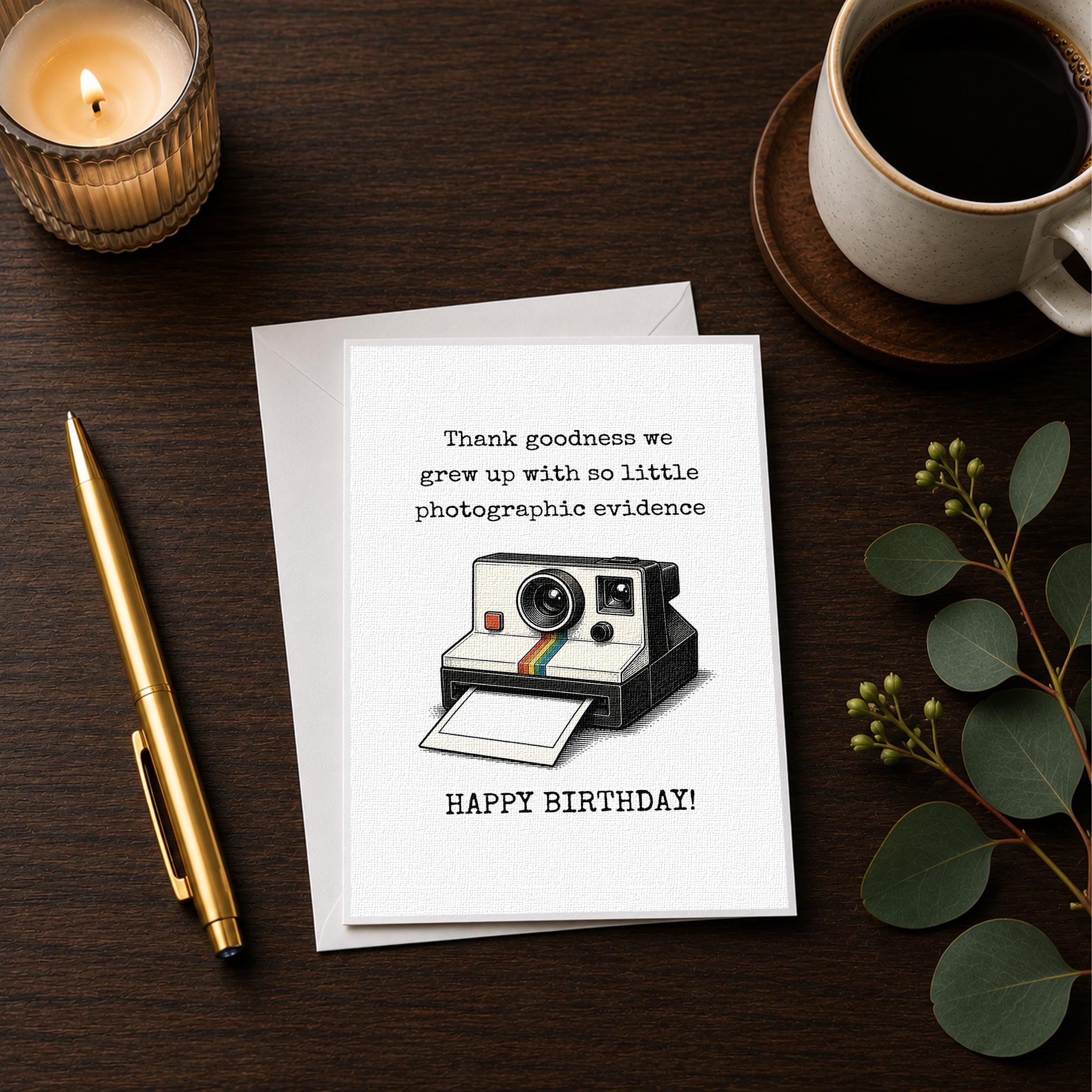 a birthday card with a camera illustration, a pen, a candle, and a cup of coffee on a wooden surface.