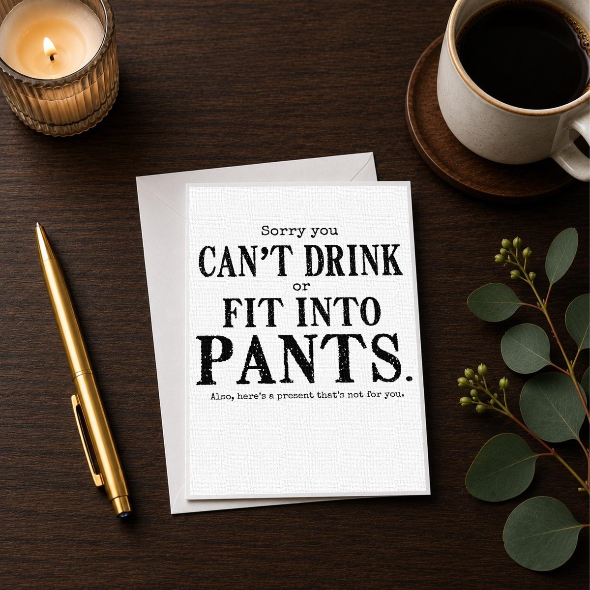 White 5-by-7 greeting card displayed upright on a white envelope with distressed black typography reading, “Sorry you can’t drink or fit into pants. Also, here’s a present that’s not for you.”