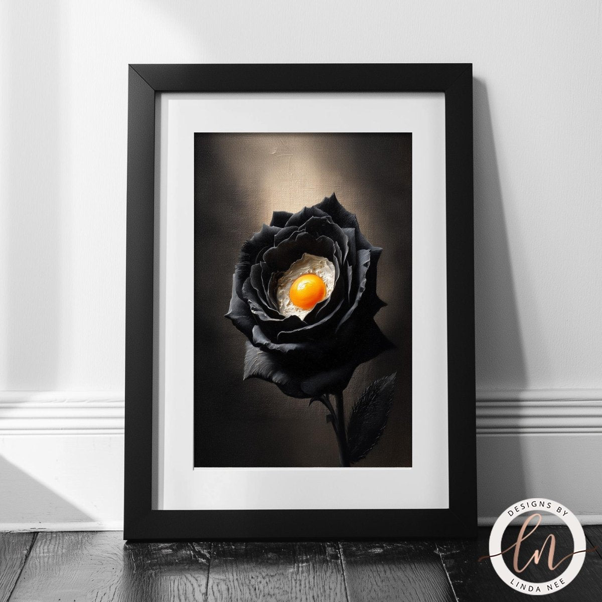 A framed photograph of a black rose with an orange center, placed on a wooden floor against a white wall.