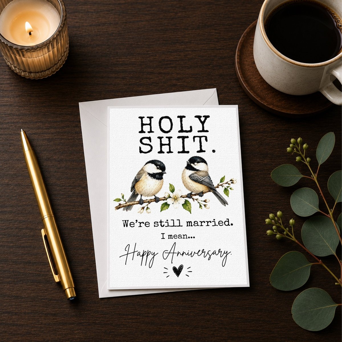 a greeting card with a humorous message, a pen, a candle, and some greenery on a wooden surface.