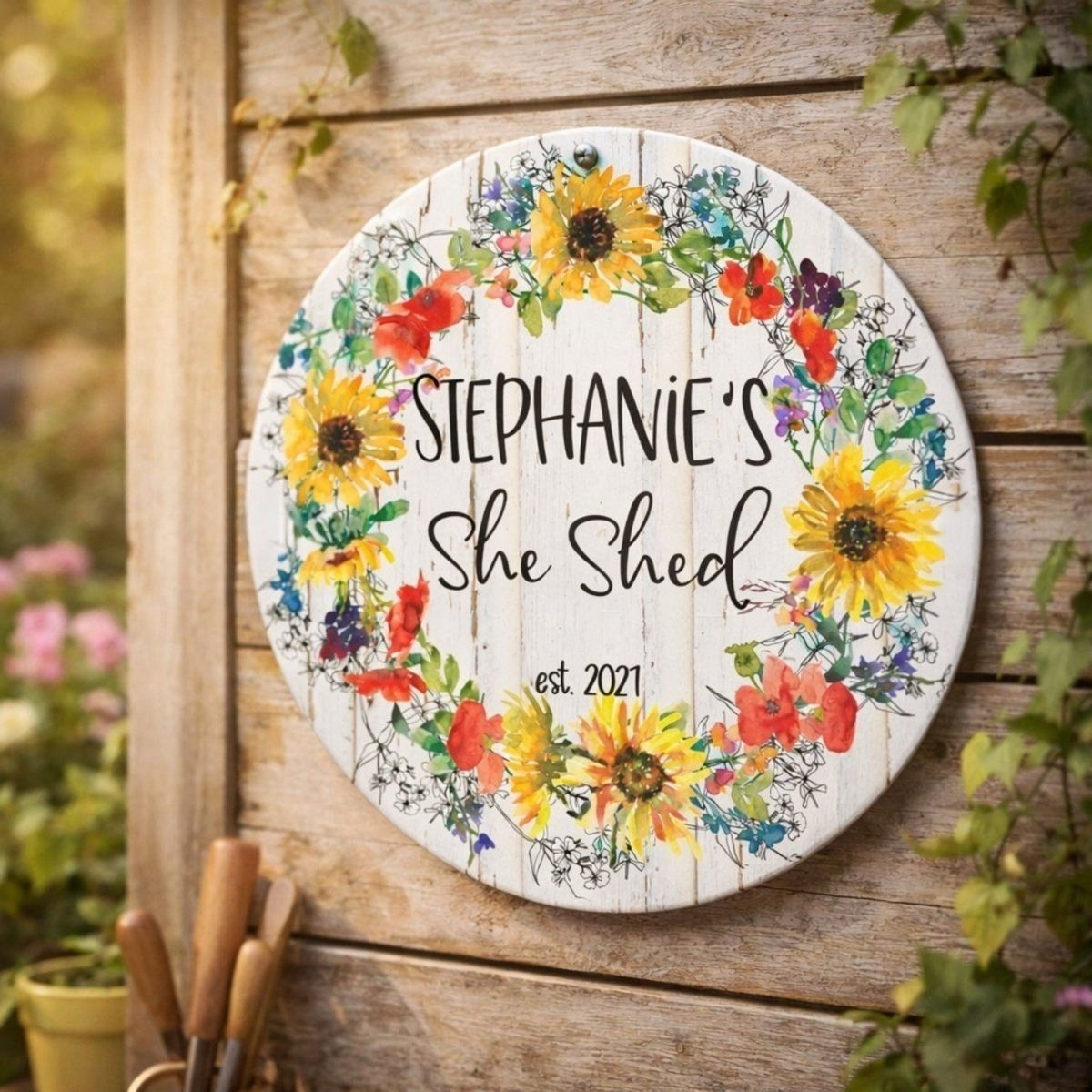 A round wooden sign with a floral wreath design hangs on a wooden wall, displaying the name "Stephanie's She Shed" and the year "est. 2021".